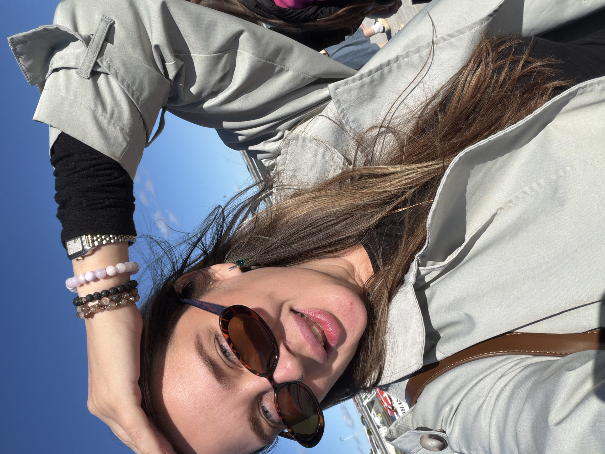 This is a selfie of a young woman with long, brown hair, taken outdoors against a bright blue sky. She is wearing a light gray trench coat over a black long-sleeved shirt. On her left wrist, she has a silver watch and three beaded bracelets, one in a light pink hue, one of black beads, and one with brown and clear beads. She is also wearing oval-shaped sunglasses with tortoise shell frames. A small green earring is visible in her left ear. Her skin appears fair and she has light makeup on. Her lips are slightly parted, and she has a gentle smile. In the background, some other people and a glimpse of what appears to be a street or market scene are visible, along with some partial text on what might be a sign or banner. The lighting suggests it is daytime, possibly late morning or early afternoon, with clear weather. The context provided places the location as Sortavala, Russia.