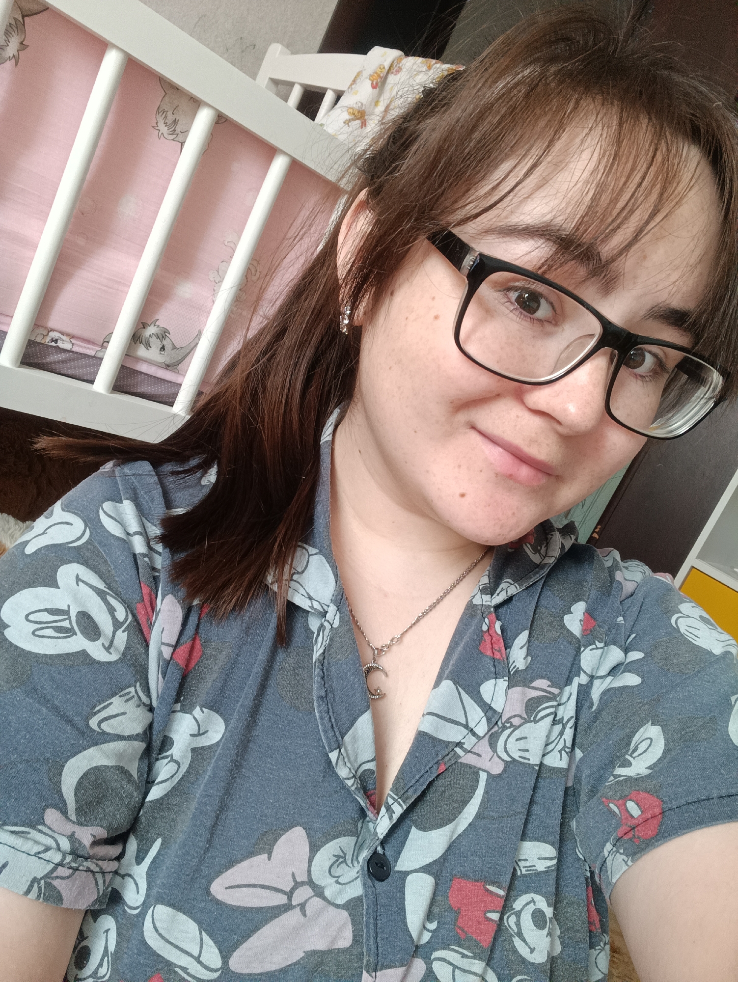 A close-up, eye-level shot features a woman with brown hair pulled back in a partial ponytail, wearing large, black-framed glasses and a blue button-down shirt patterned with cartoon characters. She has fair skin with freckles and is smiling softly at the camera. A silver necklace with a crescent moon pendant is visible around her neck. Behind her, a white crib with vertical slats and a pink interior is partially visible to the left. The location is Unknown, Unknown.