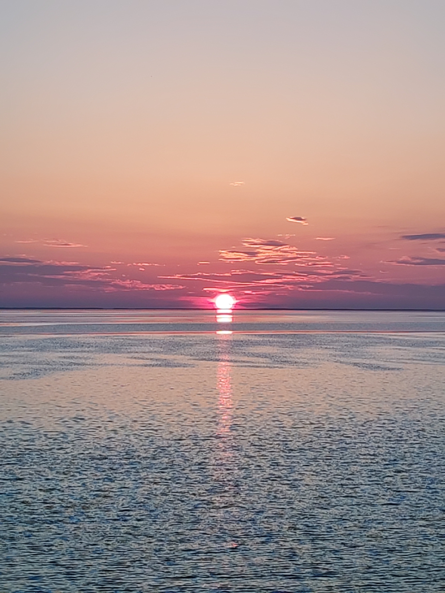 The image depicts a sunset over water. The sun is positioned near the horizon, partially obscured by clouds. The sky is colored in shades of pink, orange, and light purple. The water reflects the sunlight, creating a bright line extending towards the viewer. The water has small ripples across its surface.
Location: Unknown, Unknown.