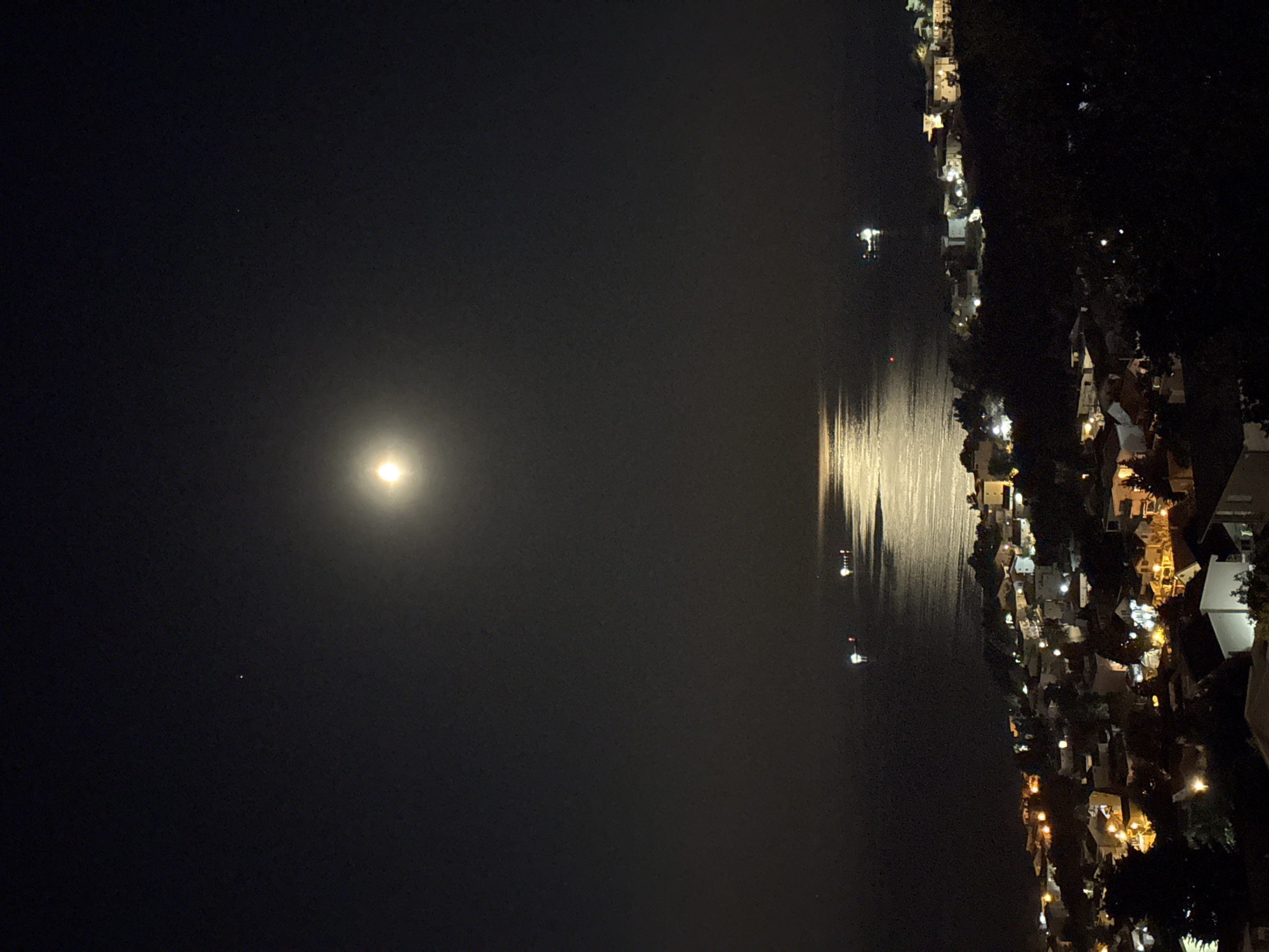 A nighttime view captures the city of Šušanj, Montenegro, oriented vertically with the dark sky dominating the left half of the frame. A bright moon is positioned in the upper-left quadrant. Its reflection creates a shimmering, vertical line of light on the water. Along the right edge is a densely packed collection of buildings and houses, illuminated with various warm-toned lights, indicative of residential areas. Additionally, there are at least four boats on the water with illuminated lights. The scene lacks any human presence.