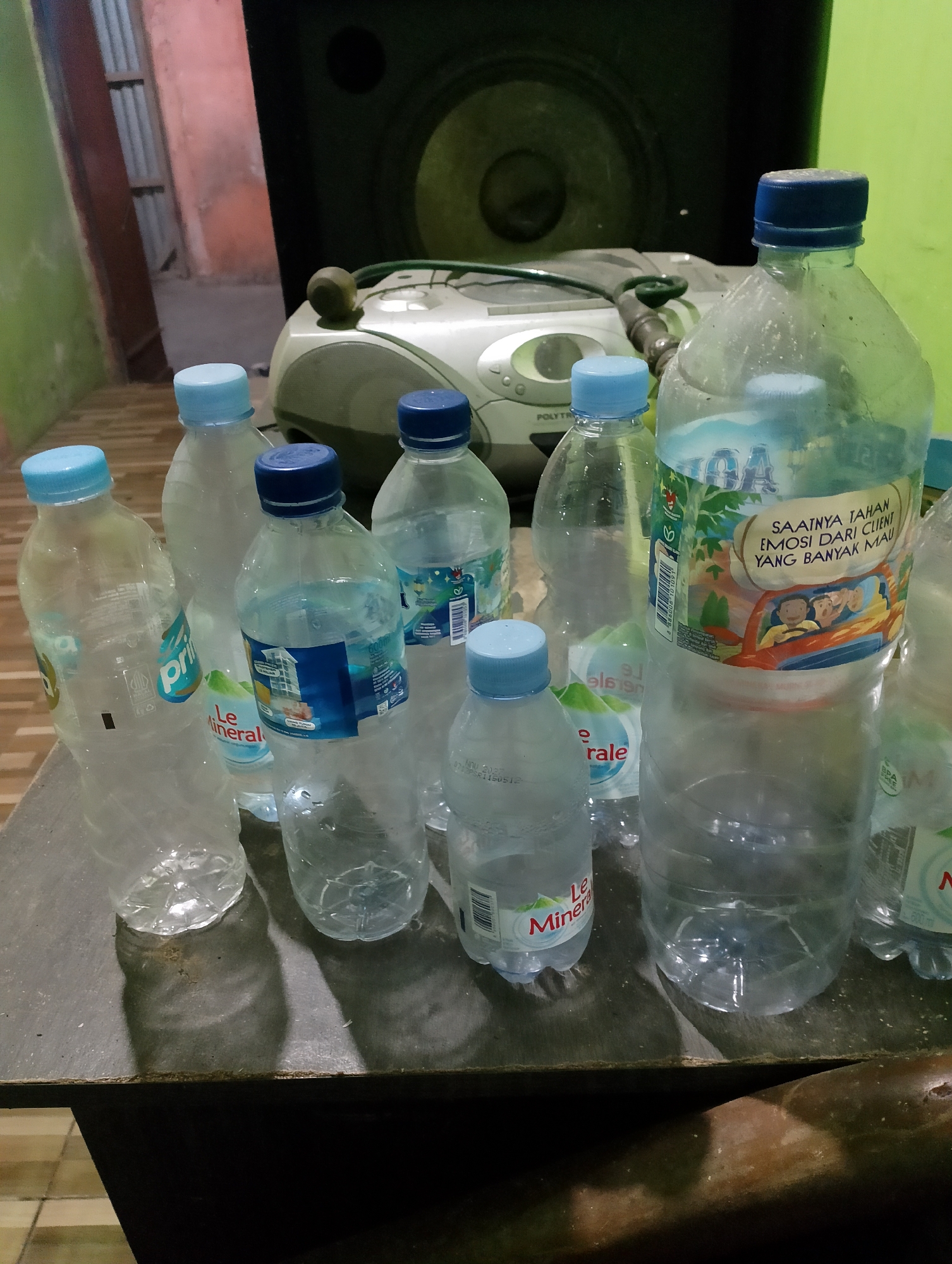 The image shows a collection of empty and partially empty plastic water bottles on a dark wooden surface. The bottles have blue caps and various labels, some indicating brands like "Le Minerale" and "AQUA". In the background, a large speaker is visible, along with a white electronic device. The environment appears to be indoors, possibly in a storage area or a casual setting, with hints of green and pink walls. The scene is dimly lit. There are no people visible in the image. The exact location in Rembang, Indonesia, is not determinable from the image itself.