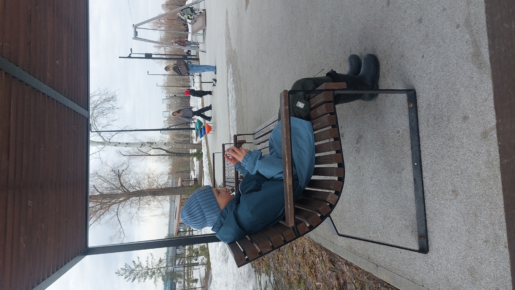 A photograph captured in Novosibirsk, Russia, shows a young person sitting on a wooden bench in what appears to be a public park or recreational area. The individual is wearing a blue winter hat and a blue jacket, facing forward. The person is looking at or interacting with a mobile device. The bench is positioned on a concrete surface. In the background, there are several people in motion and what appears to be playground equipment. The sky is overcast. A wooden structure, potentially part of a shelter or shaded area, is visible on the left side of the frame. The ground in the area contains patches of snow. The boots are black.