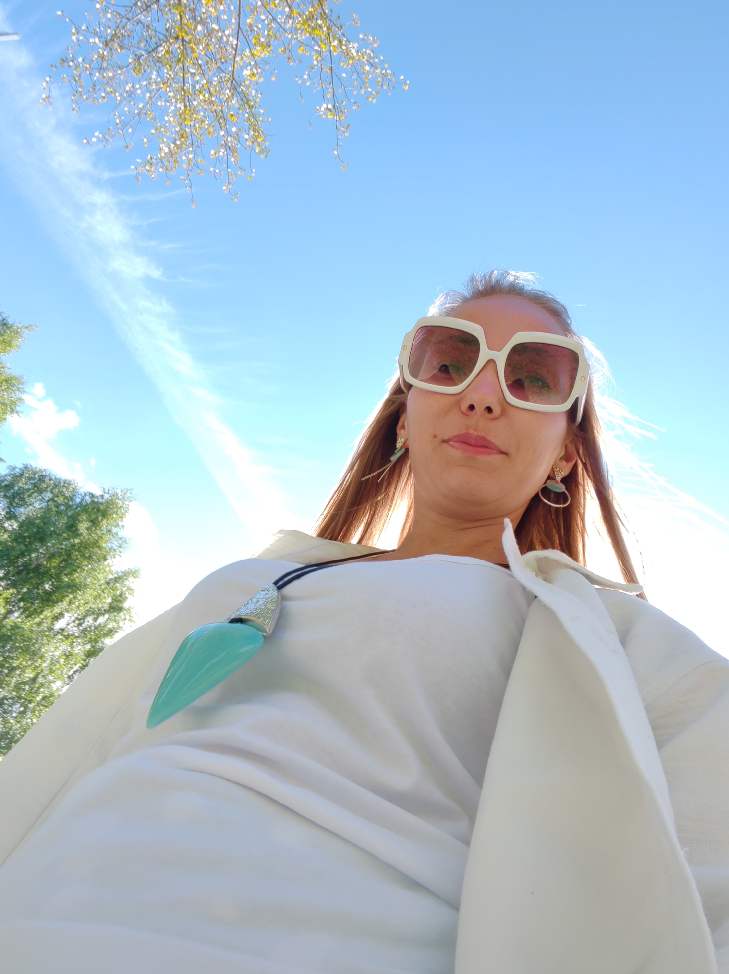 This is a low-angle, waist-up shot of a woman wearing large, white, square-framed sunglasses with pink lenses. She has long, brown hair and is wearing a white top with a white jacket draped over her shoulders. A large, teal pendant hangs from a black cord around her neck. She is also wearing elaborate, dangling earrings. The background is a bright blue sky with some wispy white clouds and sunlight filtering through the leaves of a tree in the upper left corner. There is also greenery visible on the left side of the image, suggesting an outdoor setting.
The provided information states that the location is Syktyvkar, Russia, but the image itself does not contain any specific visual cues that would confirm this. Based solely on the visual content of the image, it appears to be an outdoor scene on a sunny day.
