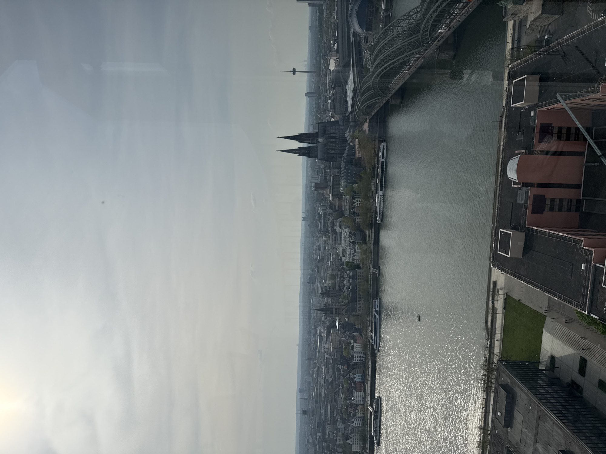The image shows an aerial view of Cologne, Germany, with the Rhine River running through the city. The Cologne Cathedral is a prominent feature, as is the Hohenzollern Bridge. Riverboats are visible on the Rhine. The scene appears to be taken from a high vantage point, likely a tall building or observation deck. The environment is overcast with a hazy sky.