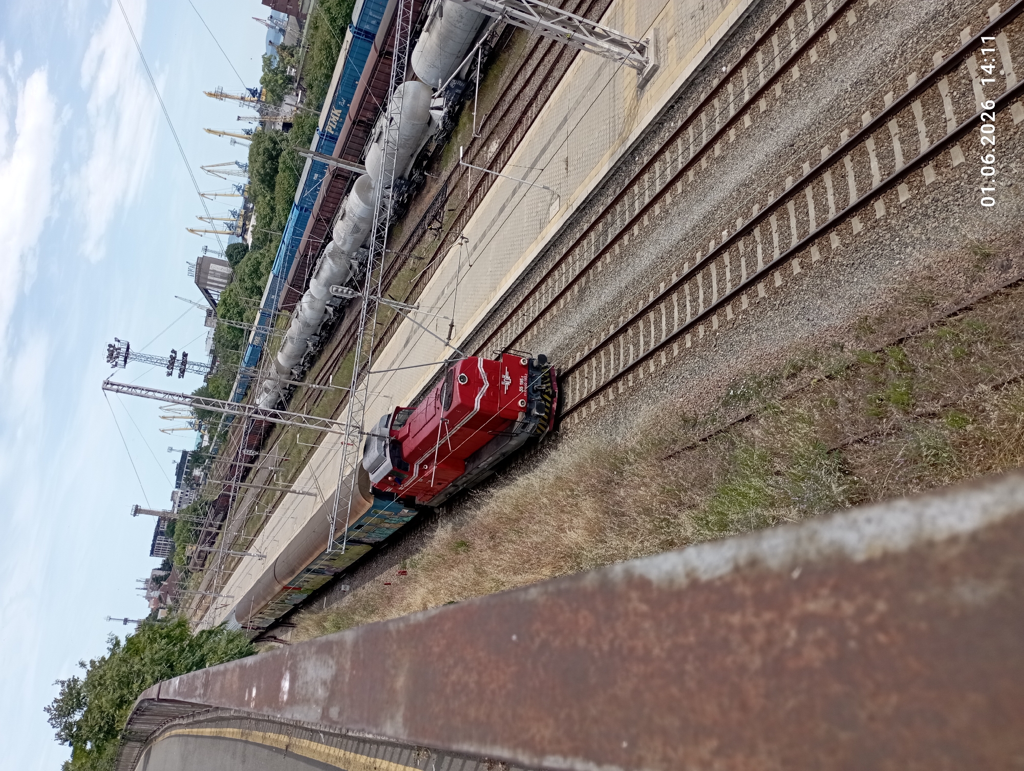 The image shows a train yard with multiple tracks and a red locomotive on one of the tracks. The photograph is taken from an elevated perspective, with a rusty railing in the foreground. In the background, there are train cars, storage tanks, and cranes, suggesting a port or industrial area. The sky is visible in the upper portion of the image. There is text "01.06.2026 14:11" in the bottom-right corner.