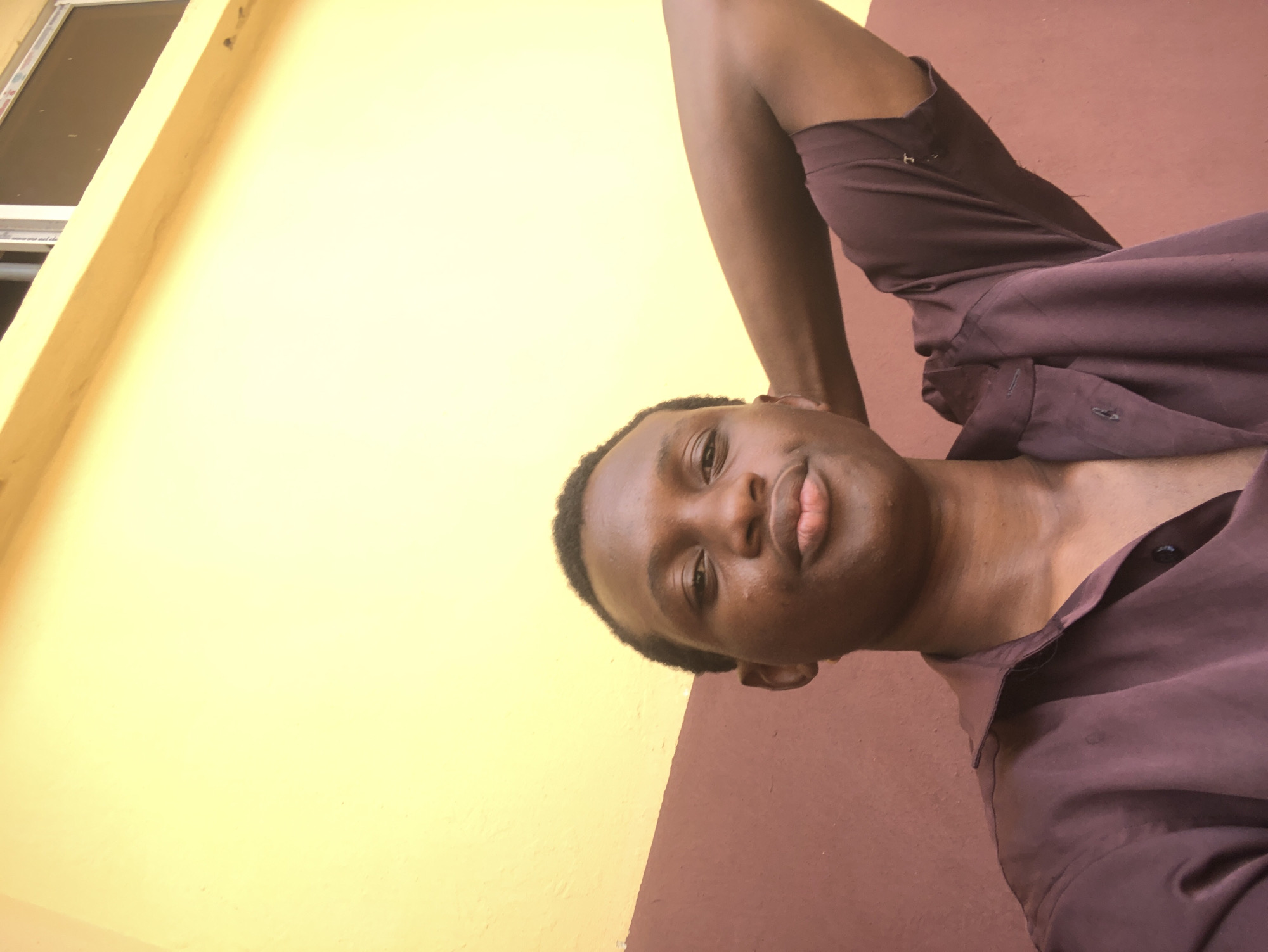 A person is taking a selfie. The person is dark-skinned, wearing a brown shirt, and has short, dark hair. The background consists of a wall that is yellow on the upper part and brown on the lower part. A window is visible in the top left corner. It is unknown if the person is located in Ukwu-Abwa, Nigeria.