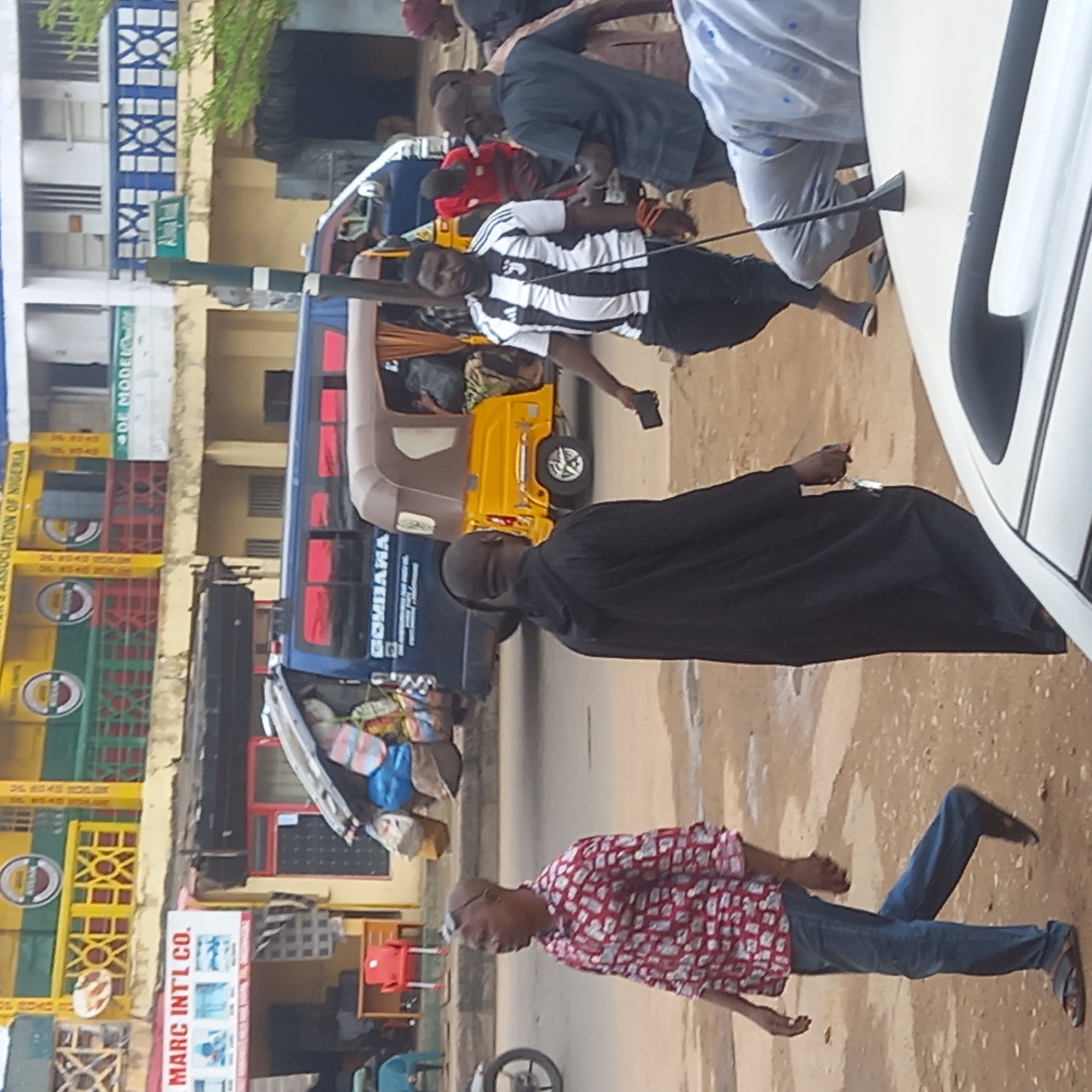 The image depicts an urban street scene in Dadi Riba, Nigeria. Several people are visible, including a man in a red and white patterned shirt and jeans walking away from the camera, another man in a black robe walking towards the right, and a man in a black and white striped shirt standing near a yellow auto rickshaw. A dark blue bus is parked behind the rickshaw. A white vehicle is partially visible on the right side of the image. The background includes multi-story buildings with signs, including one that reads "MARC INTL CO.". Another sign displays the words "GOMBARA" on the side of the bus.