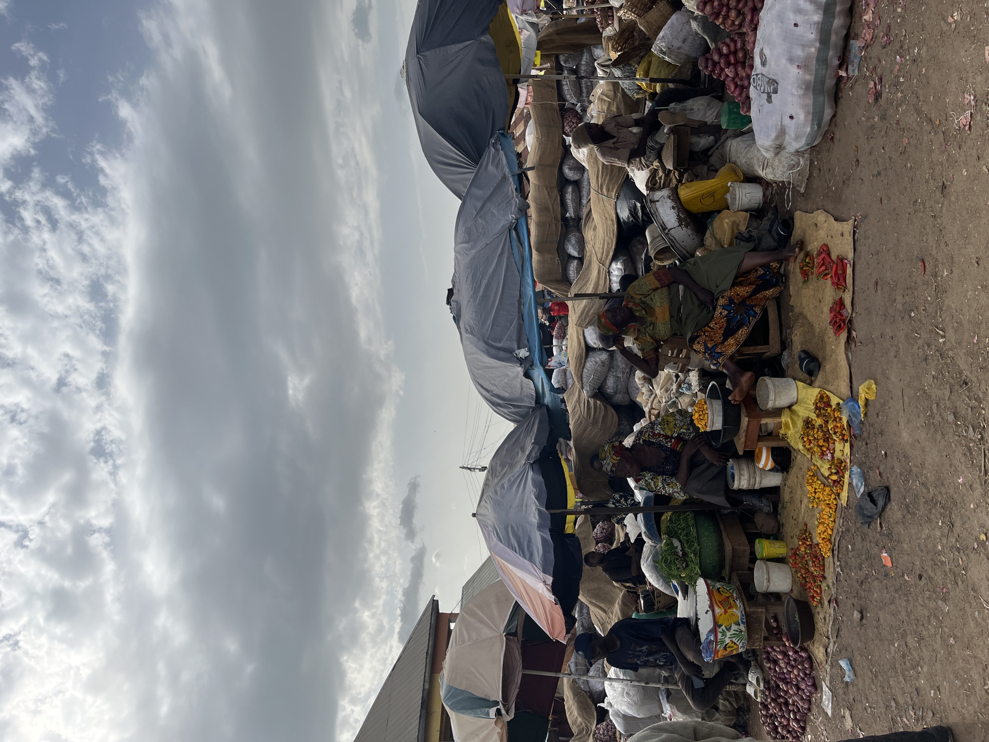 The image depicts a bustling outdoor market scene in Rafin Par, Nigeria, under an overcast sky. The perspective is vertical, creating a sense of depth and scale. In the foreground, several individuals are engaged in market activities. A woman sits on a mat, tending to a display of red and yellow fruits, possibly tomatoes or peppers, and some onions. She is dressed in a colorful patterned skirt and head wrap. Beside her, various containers, including buckets and a yellow plastic container, hold produce. Further back, more people are visible, some tending to stalls laden with sacks of produce, including onions and what appear to be grains. Large umbrellas are erected to provide shade over the stalls. The ground is dusty and appears to be a dirt path. The overall atmosphere suggests a busy commercial hub, with a focus on agricultural products. The time of day is likely daytime, given the light, although the cloudy conditions obscure direct sunlight. No discernible text is visible in the image.