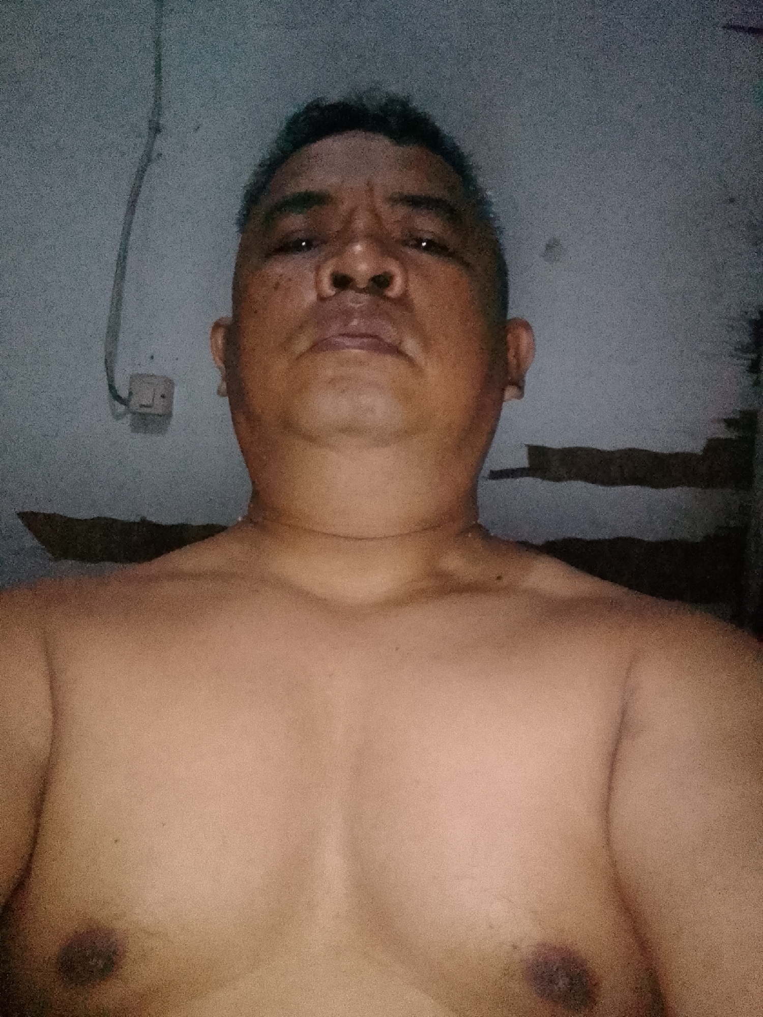 A medium shot, taken from a slightly low angle, shows a shirtless man from the chest up. He has short, graying hair and is looking directly at the camera with a neutral expression. His skin is tan, and he has a pronounced neck. The background is a dimly lit, textured wall, likely indoors, with a white electrical outlet and a wire visible on the left. A dark, horizontal band runs across the middle of the background. The image quality is low, with significant grain and noise. The specific location within Gunungmasigit, Indonesia, is not identifiable from the visual information provided.
