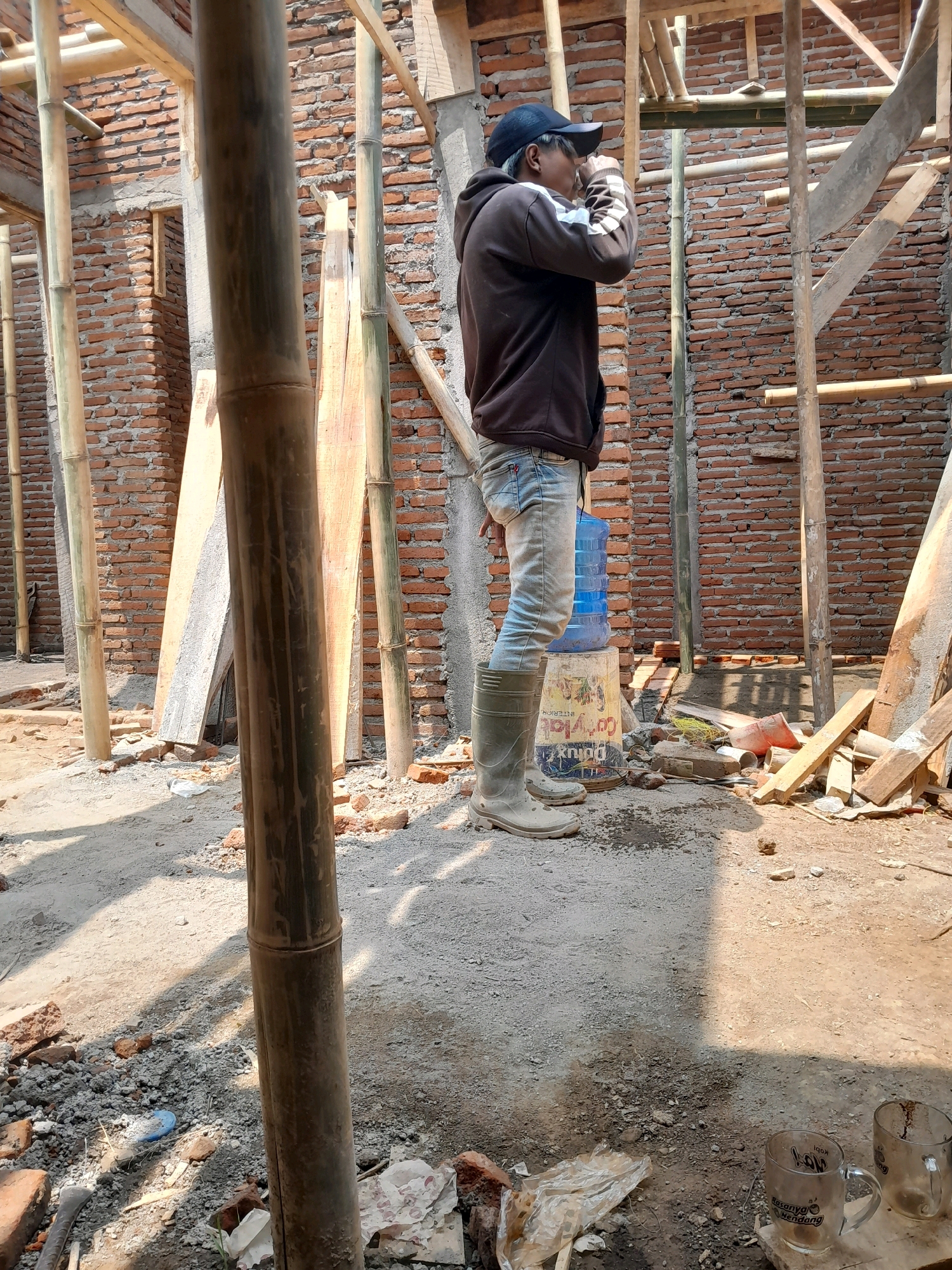 A man wearing a dark hooded sweatshirt, light blue jeans, and tall grey rubber boots is standing at a construction site. He is holding a cup to his mouth, appearing to drink. Behind him, a blue water cooler bottle sits on a cylindrical container. The construction site features unfinished brick walls, bamboo scaffolding, and wooden planks. The ground is covered in dirt and rubble. In the foreground, two glass mugs are visible on the ground. The scene is illuminated by natural sunlight, casting shadows. The man's posture and attire suggest he may be a worker taking a break. The location is identified as Karangduren, Indonesia.