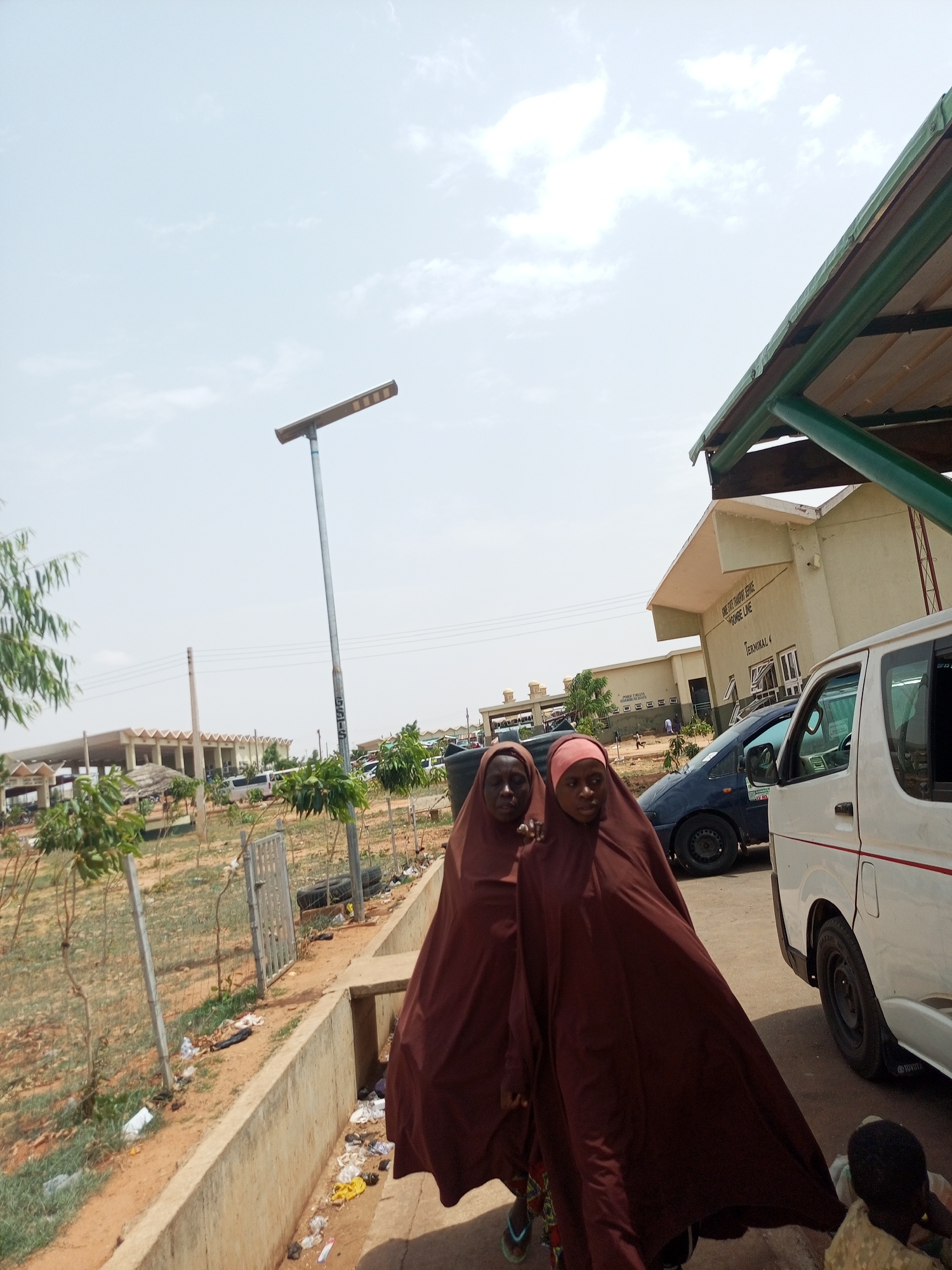 Two women wearing matching brown hijabs walk on a walkway in front of what appears to be the Gombe Line Transport Service Highgate Line Terminal 4 in Gombe, Nigeria. A white passenger van and a dark-colored sedan are parked nearby. A solar-powered street light stands tall in the background. The walkway is bordered by a concrete curb and a wire fence. Trash litters the ground. In the background, a low building with a thatched roof stands amidst sparse greenery. The sky is light blue with scattered clouds.
