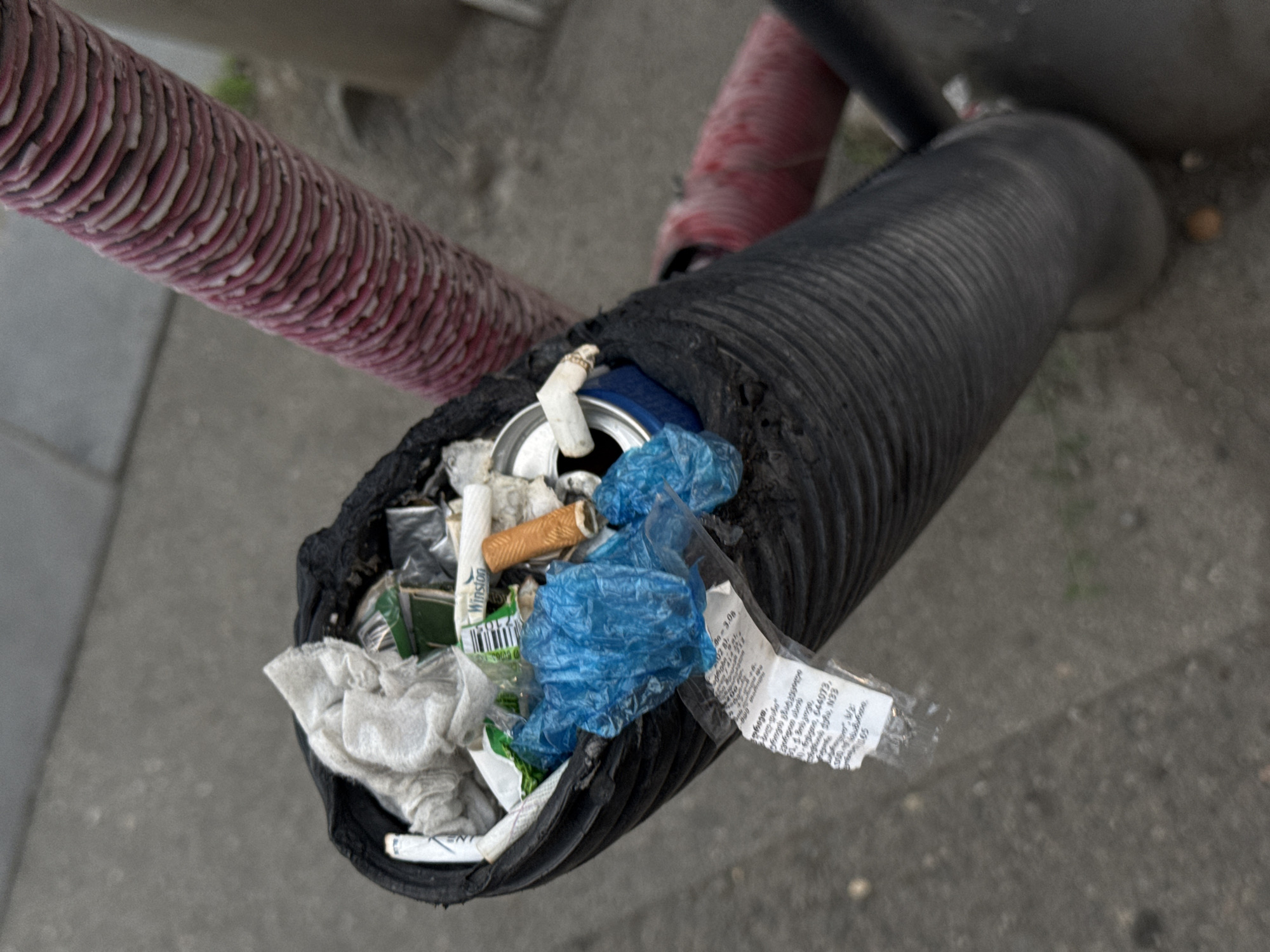 The image shows a close-up, high-angle view of a black, corrugated pipe stuffed with garbage. The garbage includes crumpled white paper or cloth, plastic bags, cigarette butts, a metal can, and other debris. A label with text, possibly in Georgian, is partially visible and hanging out of the pipe. In the background, a pink corrugated tube runs diagonally across the frame, and the ground appears to be a concrete or asphalt surface. The lighting suggests an overcast day. The scene is an outdoor urban environment, consistent with a street in Tbilisi, Georgia. There are no people visible in the image. The focus is on the discarded items within the pipe, suggesting a public area where waste has accumulated.