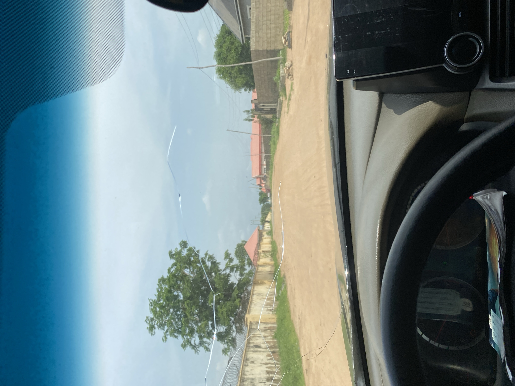 The image shows a view from inside a car looking out through the cracked windshield onto a dusty road in a residential area. The road is unpaved and appears to be in a rural or semi-urban setting. On the left side of the road, there is a long, beige wall with barbed wire on top, followed by a large green tree. Further down the road, several houses with orange roofs are visible. On the right side of the road, there are more houses constructed with concrete blocks, and utility poles with wires. The sky is cloudy, suggesting it might be overcast or a hazy day. The interior of the car is visible in the foreground, including the steering wheel and a portion of the dashboard with a digital display. There are no people visible in the image. The overall scene depicts a typical street in a developing area, possibly Howayi, Nigeria, characterized by simple architecture, unpaved roads, and visible infrastructure.