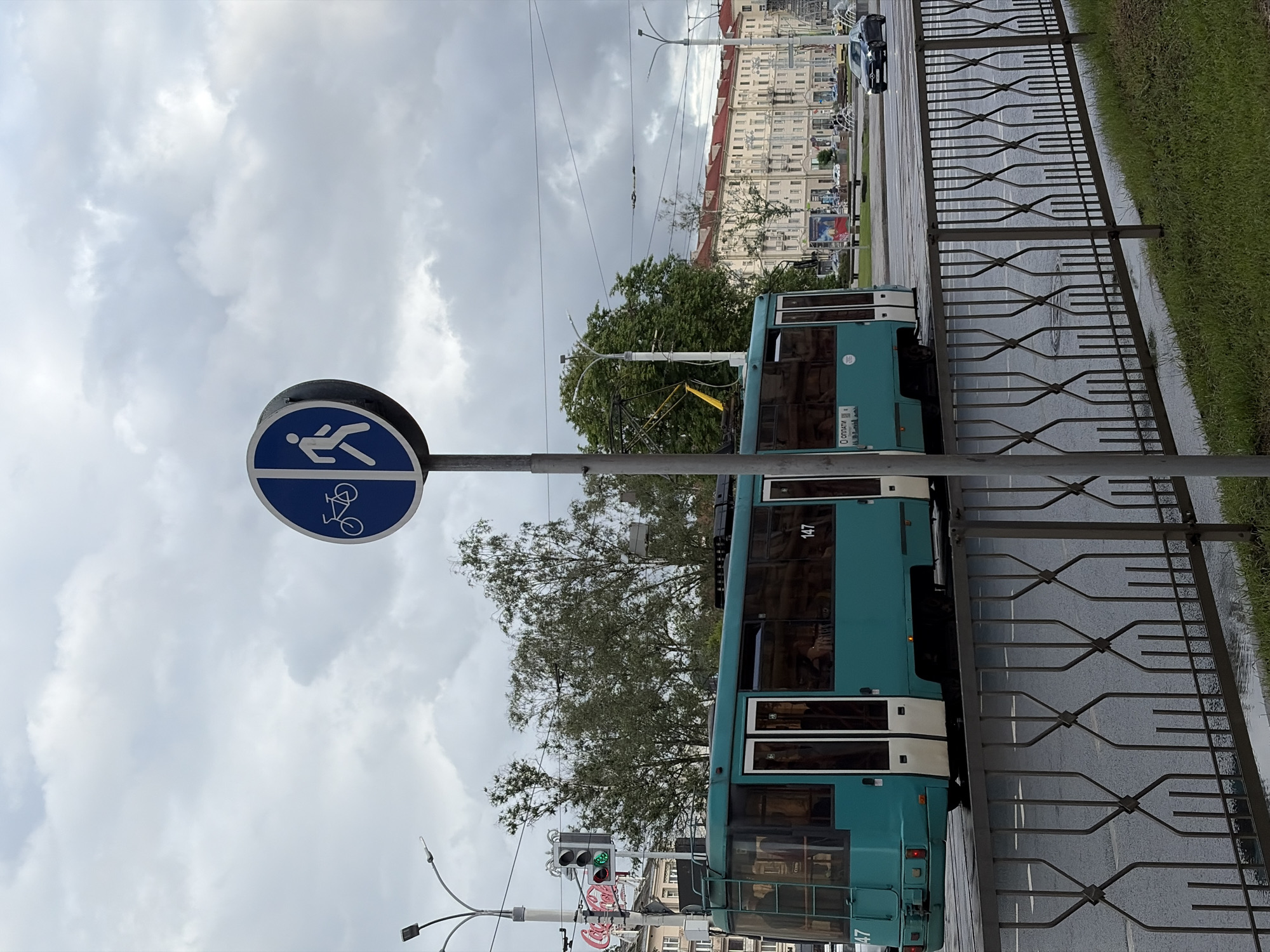 The image shows a street scene in Minsk, Belarus, on a cloudy day, possibly in the late afternoon or early evening. A teal-colored tram with white trim is the most prominent object, positioned parallel to a metal fence that separates the tram tracks from a grassy verge. The tram has the number "147" visible on its side. A traffic light, showing green, is also visible.
To the left of the tram, a blue circular sign with white pictograms indicates a shared path for pedestrians and cyclists. The sign features a silhouette of a person walking and a bicycle. Overhead, a network of power lines crisscrosses the sky, suggesting the presence of the tram's electrical system. In the background, a multi-story building with a red roof is visible, characteristic of urban architecture. The street appears wet, indicating recent rain.
The overall mood is subdued due to the overcast weather. The scene is a typical urban streetscape, highlighting public transportation and shared pedestrian and cycling infrastructure. There is a small amount of Cyrillic text visible on the tram, likely related to its route or destination.