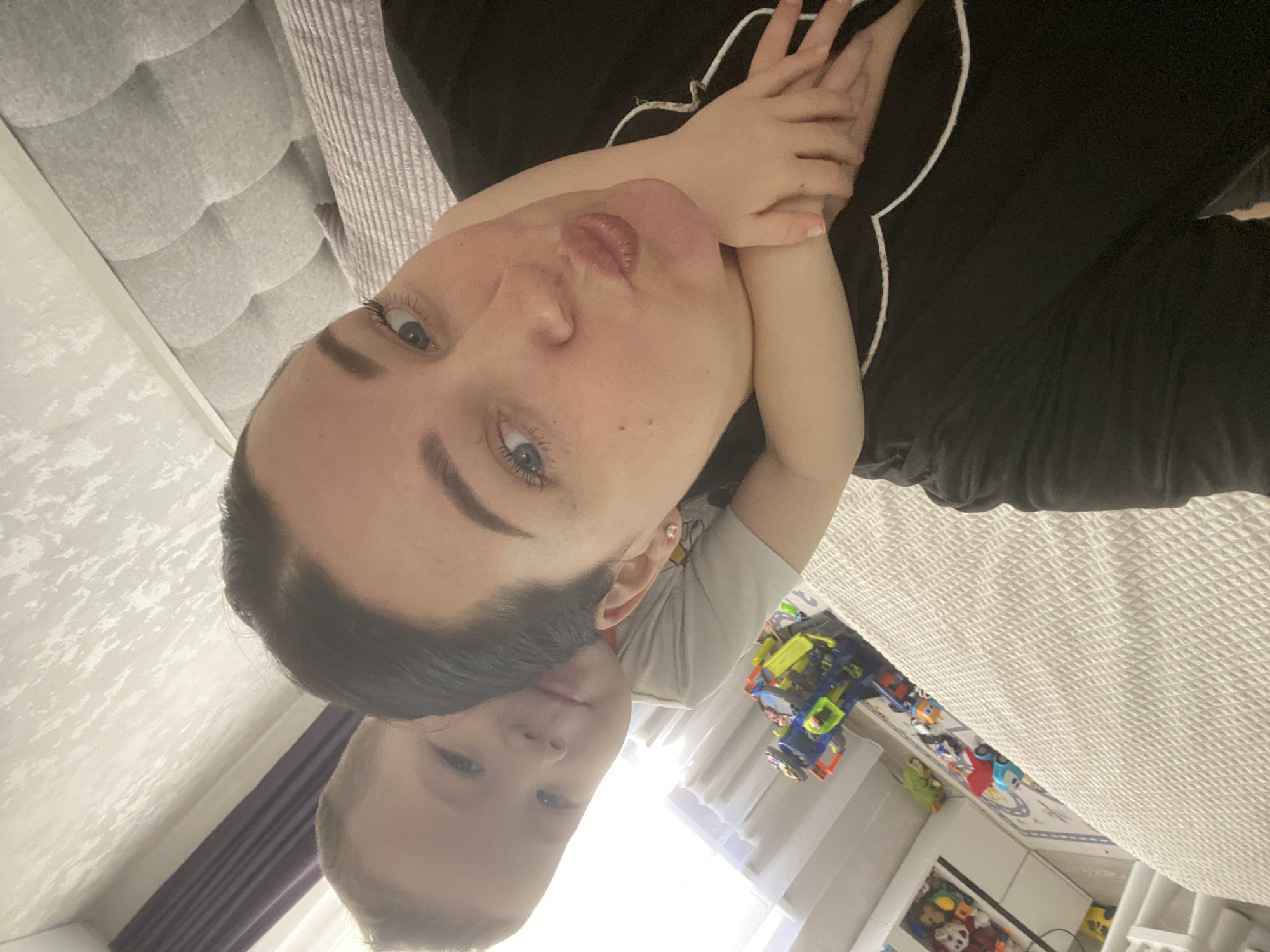 A woman and child are lying on a bed. The woman is wearing a black shirt with white piping and has dark hair. The child is embracing the woman's neck with their arms. The child is wearing a gray shirt. Toys are scattered on the floor. The location is potentially a private residence. There is no visual information available to determine the location within the city of Sochi, Russia.