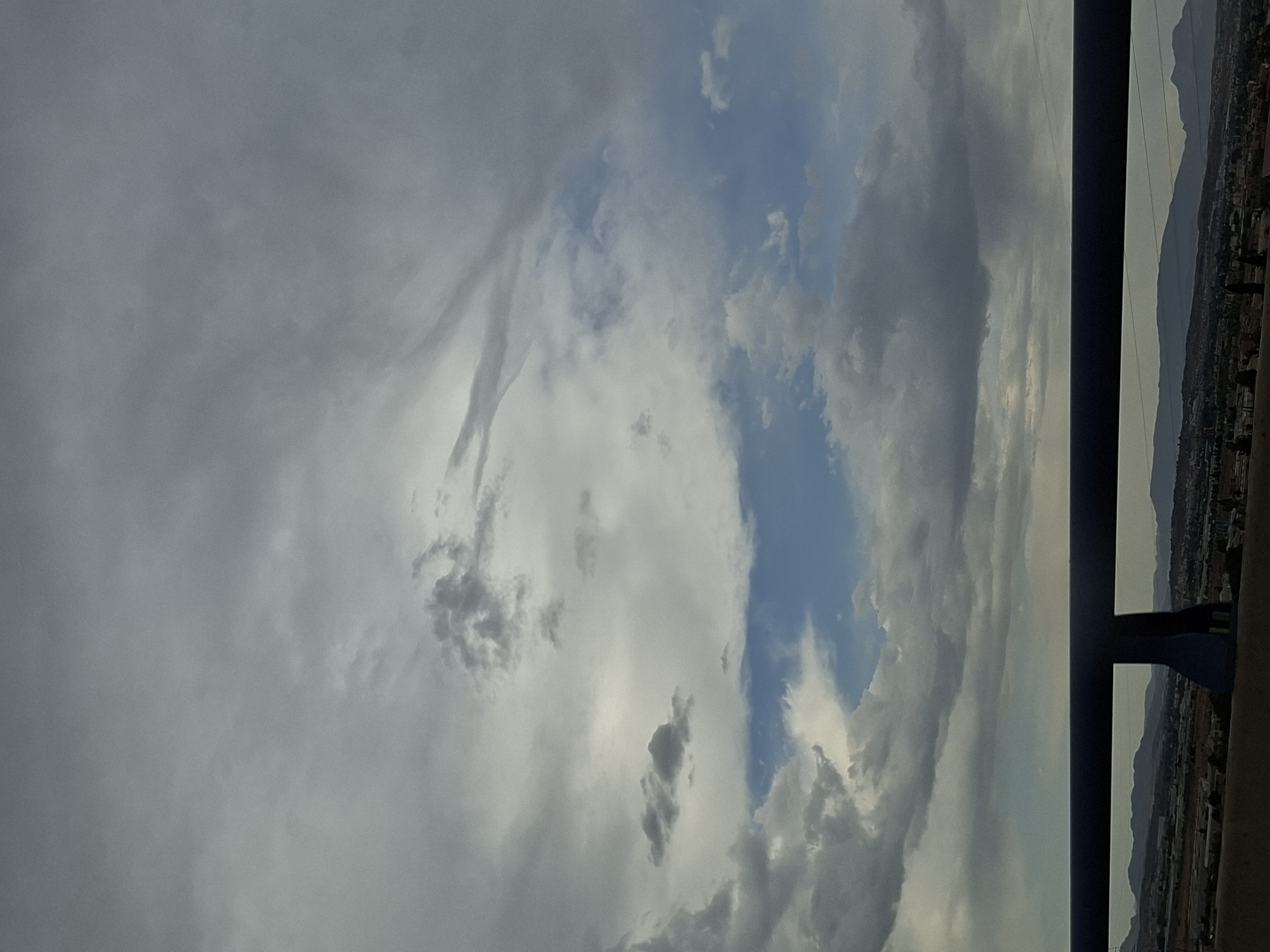 The image shows a cloudy sky over a landscape. In the foreground, a dark blue railing runs horizontally across the frame, obscuring the lower part of the scene. Beyond the railing, a wide expanse of mountains and a town can be seen. The sky is filled with various cloud formations, ranging from dense gray clouds to lighter, wispy clouds. There are also patches of blue sky visible, suggesting a break in the clouds. The overall impression is one of an overcast day with a hint of sunshine peeking through. The location is identified as San José De La Montaña, Spain. There are no people or animals visible in the image.