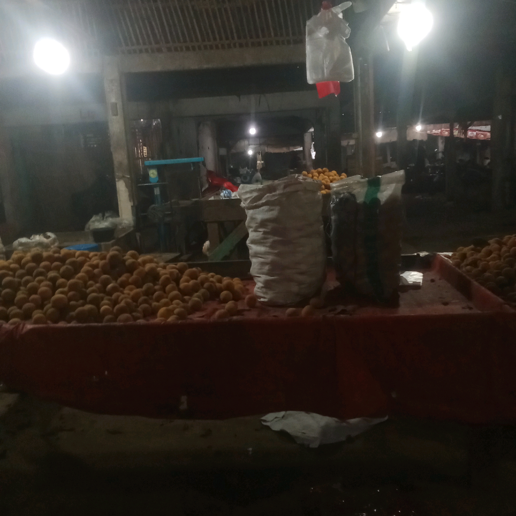 A market stall displays a large pile of small, round, orange fruits, possibly tangerines or apricots, covered by a red tarp. To the left, white and green bags are visible, and to the right, more of the same orange fruits are piled up. In the background, a tarp is hanging, and several light bulbs illuminate the scene. There are no people visible in the image. The setting appears to be a covered market or roadside stall.