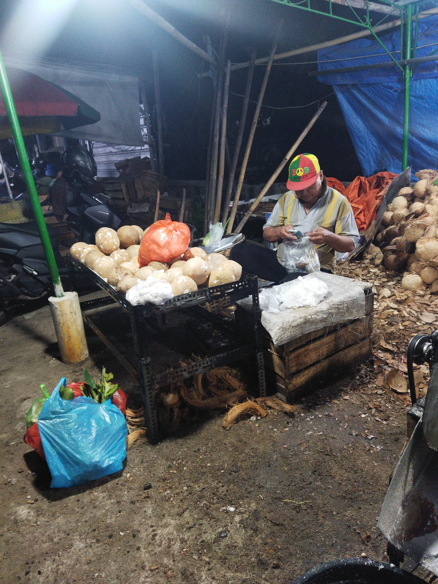 A man wearing a red, green, and yellow cap sits at a makeshift stall at night, preparing food. He is surrounded by a large quantity of coconuts, some already husked, and piles of coconut husks. In front of him is a metal rack laden with more coconuts, and a bright orange plastic bag filled with what appears to be more produce. To his left, a blue plastic bag holds green leafy vegetables. Behind him, a blue tarp and an umbrella suggest a covered outdoor market. A motorcycle is parked nearby. The ground is dirt and debris, typical of an outdoor market in Semarang, Indonesia. The man is focused on his task, and the scene appears to be a typical night market operation.