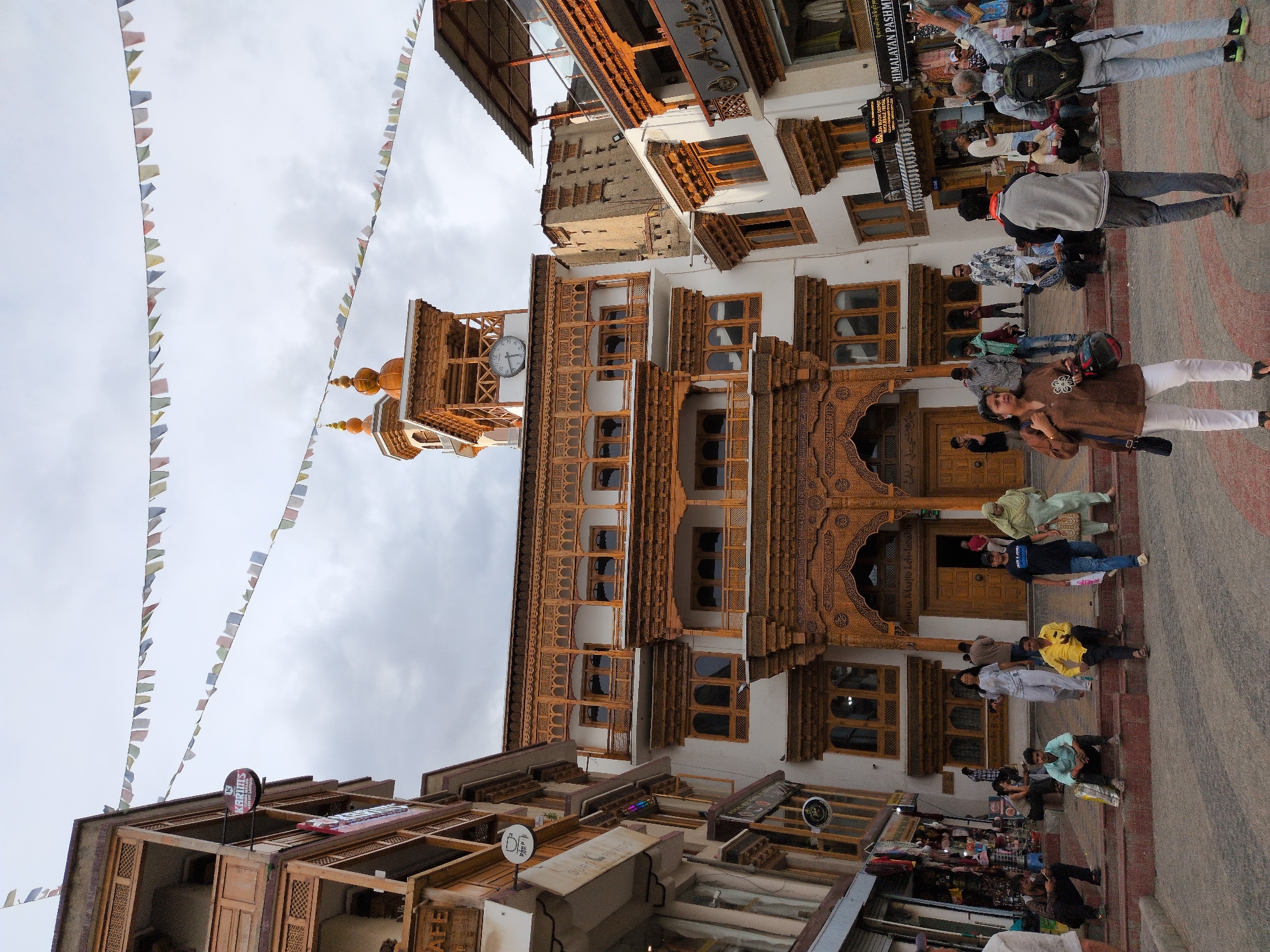 The image depicts a street scene in the city of Leh, India, likely in front of the Jamia Masjid. The architecture features traditional Ladakhi styles with intricately carved wooden balconies and facades, painted white and adorned with golden embellishments. Prayer flags are strung across the sky. A prominent clock tower with a clock face is visible atop one of the buildings. Several people are present in the scene, some seated on steps leading up to a building, while others are walking or standing. They are dressed in casual clothing. Shops and stalls line the street, with some displaying merchandise. The ground is paved with stone. The sky is cloudy, suggesting an overcast day.