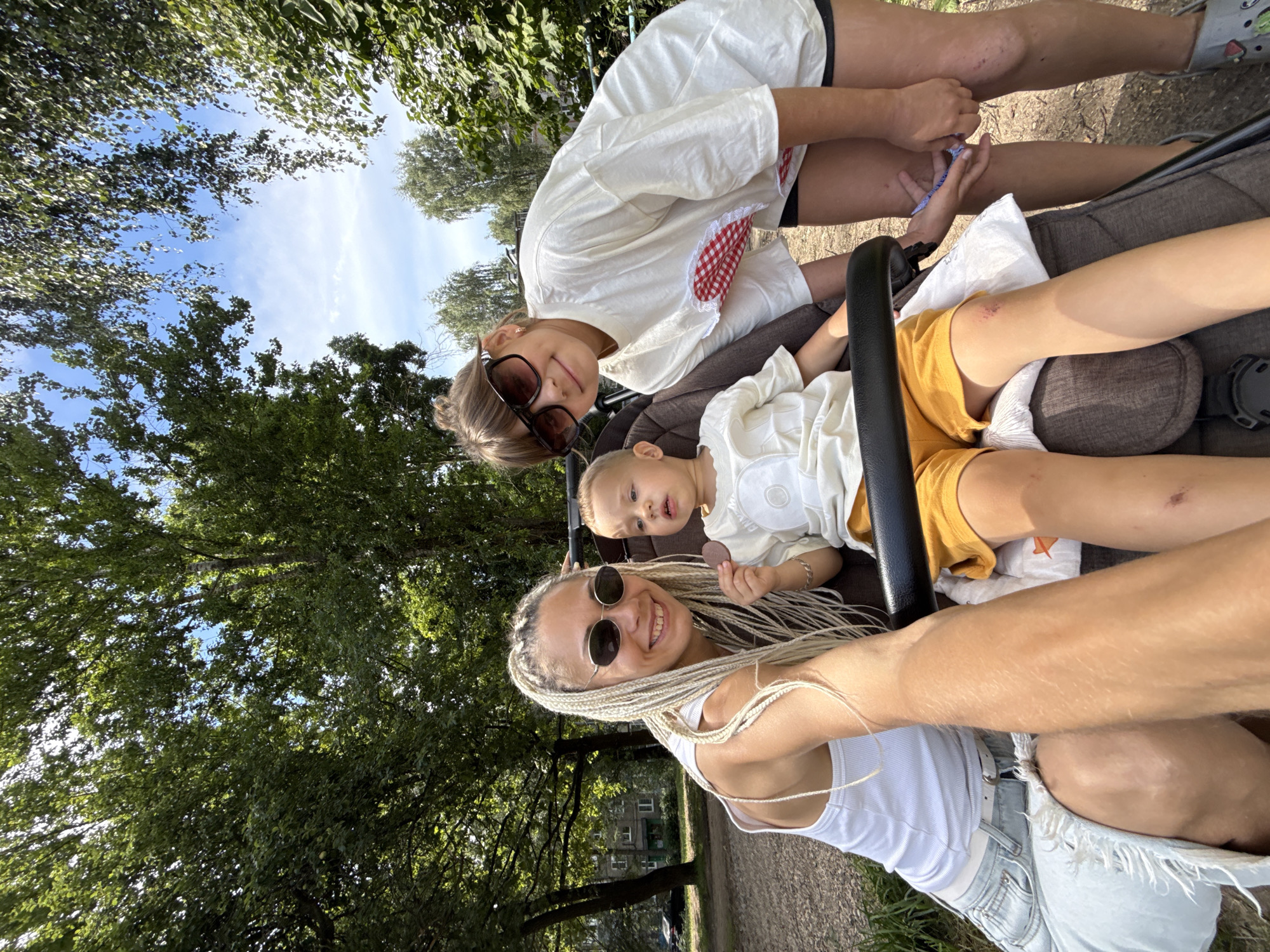 In Rusanovo, Russia, on a bright, sunny day, a woman with blonde braids and sunglasses smiles at the camera, holding a stroller with a young child. The child, wearing a white shirt and yellow shorts, sits in the stroller and holds a circular object. A teenage girl in sunglasses and a white t-shirt leans in towards the stroller, also looking at the camera. The background features lush green trees and a blue sky with scattered clouds, suggesting an outdoor park or residential area. The woman is wearing a white tank top and denim skirt, and the teenage girl is wearing a white t-shirt and black shorts. The child has a small abrasion on their left knee.