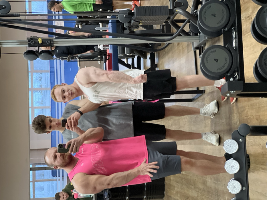 The image displays a gymnasium scene in Chaikovsky, Russia, captured as a reflection in a large mirror. In the foreground, three men are visible. On the left, a muscular man with a beard, wearing a bright pink sleeveless top and grey shorts, holds a dark-colored smartphone to take a mirror selfie. Behind him and slightly to his right, a taller man with dark hair, in a grey sleeveless shirt and black shorts, also holds a smartphone directed at the mirror. To the right of the central figure, a third man with red hair, wearing a white sleeveless top and black shorts, poses with his right arm flexed towards the mirror.
In the immediate foreground, a black rack holds multiple dumbbells, some bearing the "TECHNOGYM" brand. The background features various other gym equipment, including a prominent cable machine on the right side. Reflections in the mirror reveal additional individuals within the gym, including a man in a bright green long-sleeved shirt and black shorts further to the right, and another man partially visible to the left. The gym floor is composed of light-toned wooden or laminate material. Windows are visible along the upper part of the far wall in the reflection, suggesting natural light, with a blue stripe running horizontally beneath them.