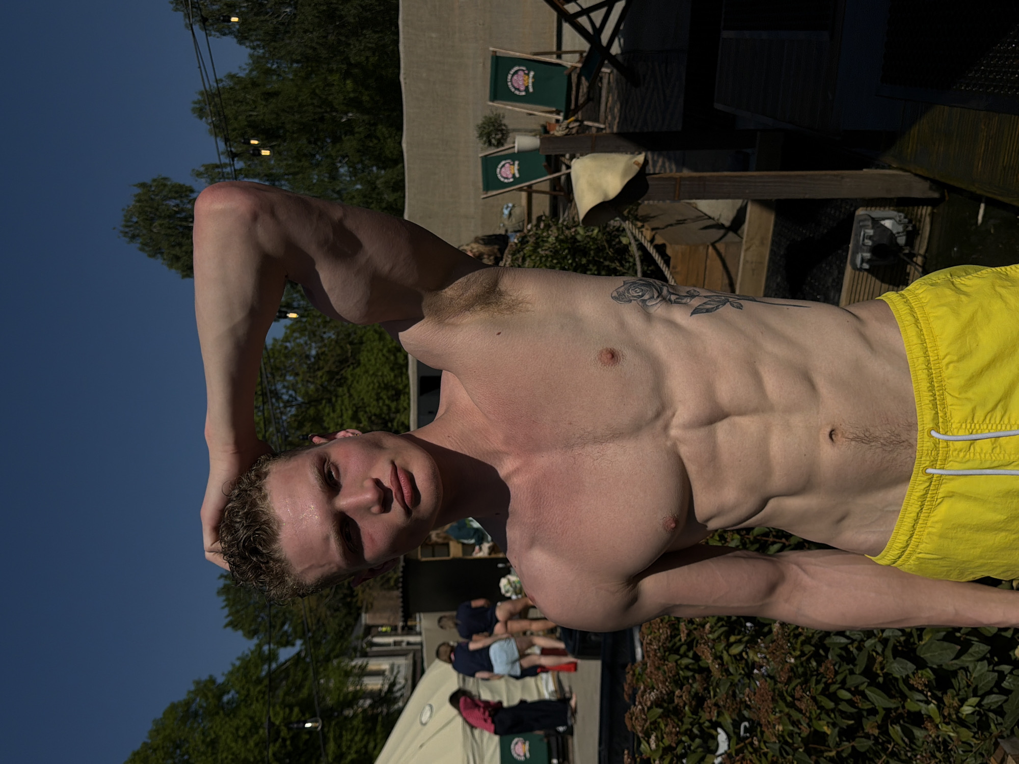 A man with a muscular physique, wearing yellow swim trunks, stands outdoors. He has his right arm raised, hand behind his head. His torso is bare, revealing defined abdominal muscles and a tattoo on his side. Behind him is an outdoor setting with people, a tent, and shrubbery. In the background, there are chairs and signs, possibly indicating a public space. The location appears to be an urban area with trees and buildings visible in the distance, potentially in the city of London, United Kingdom.