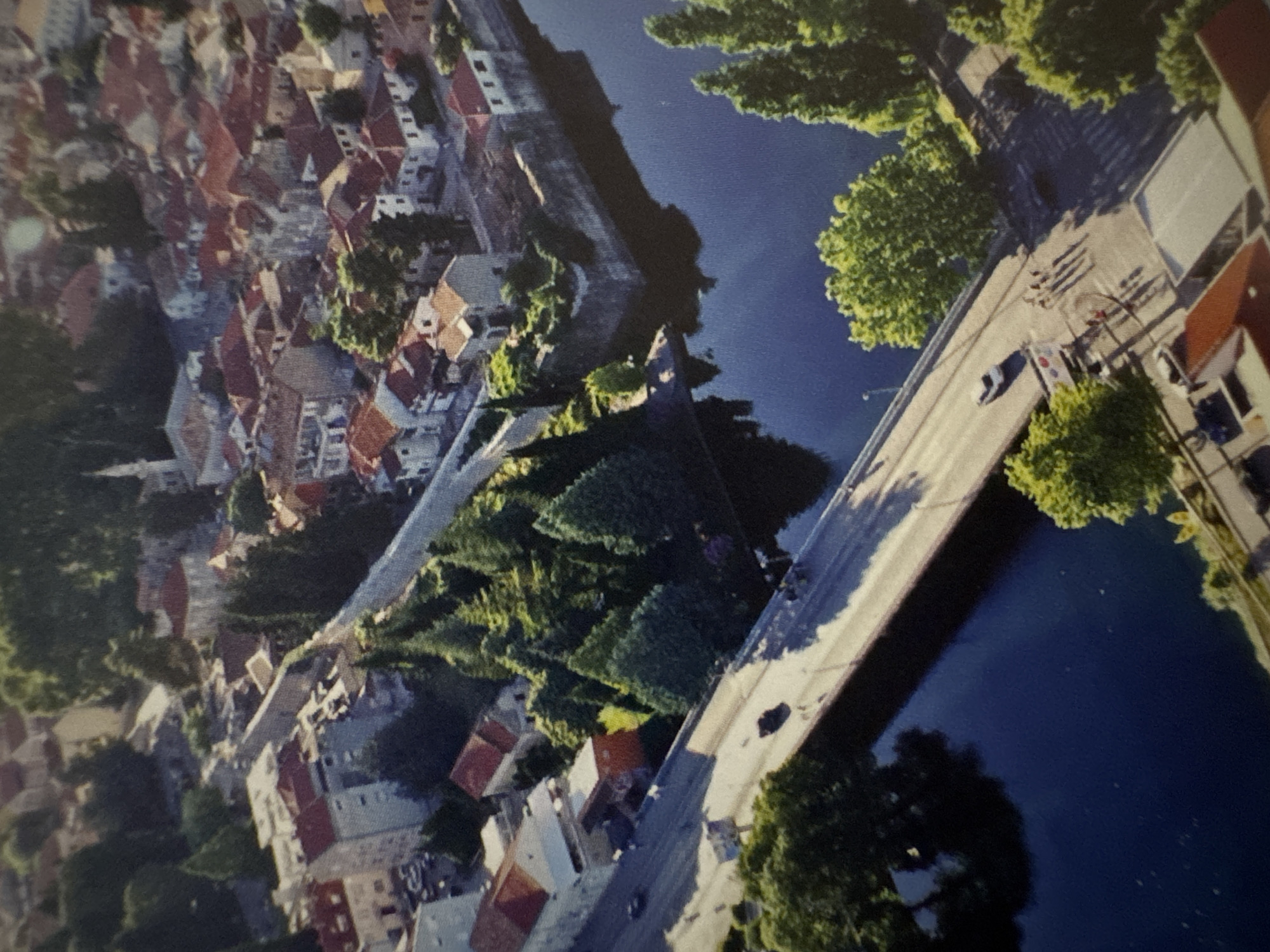 An aerial perspective depicts a city built along a wide, dark blue river under clear, sunny skies. The river runs from the upper left to the lower right, bisected by a prominent multi-lane stone bridge.
On the left bank, a series of light-colored buildings with distinct reddish-brown tiled roofs line a street running parallel to the water. Behind these structures, an ancient-looking stone wall, possibly part of a fortification, meanders through dense dark green coniferous trees. A few vehicles are visible on the street.
The right bank features a more densely packed urban area, characterized by numerous buildings with similar reddish-brown tiled roofs. A notable white building with a tall, slender minaret or spire stands prominently in the upper right quadrant, surrounded by other residential or commercial structures. A reinforced stone embankment forms a corner along the river's edge on this side.
The wide stone bridge, spanning the river, carries vehicle traffic and pedestrians. At least two white cars and one dark-colored car are observed on the bridge's lanes. A group of approximately four to five pedestrians is visible on the sidewalk on the right side of the bridge. Lush green deciduous trees line the approaches and parts of the bridge. The strong sunlight casts prominent shadows from the bridge onto the dark water and from the buildings and trees across the scene.