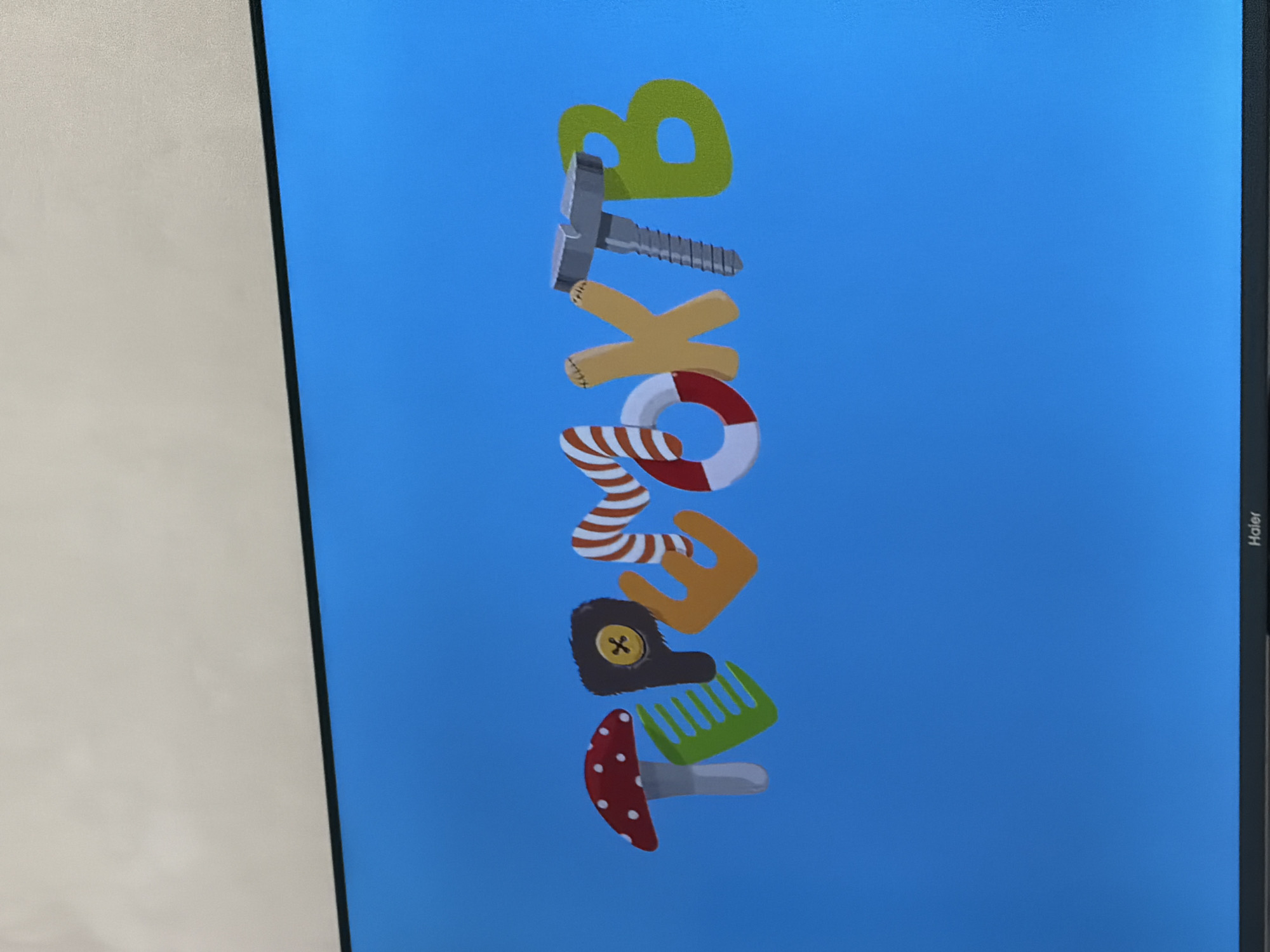 The image shows a television screen displaying the word "ТРЕОЖТВ" in stylized, colorful letters. The letters are designed to look like various objects: 'Т' resembles a mushroom, 'Р' is a green comb, 'Е' is a fuzzy brown object with a yellow button, 'О' is a life preserver, 'Ж' is a light brown letter with stitching, 'Т' is a silver screw, and 'В' is a green letter. The background of the screen is a solid blue color. The bottom of the television screen has a black bezel with the word "Haier" printed on it. The context of Krasnyi Krym, Russia is not visually represented in the image.
