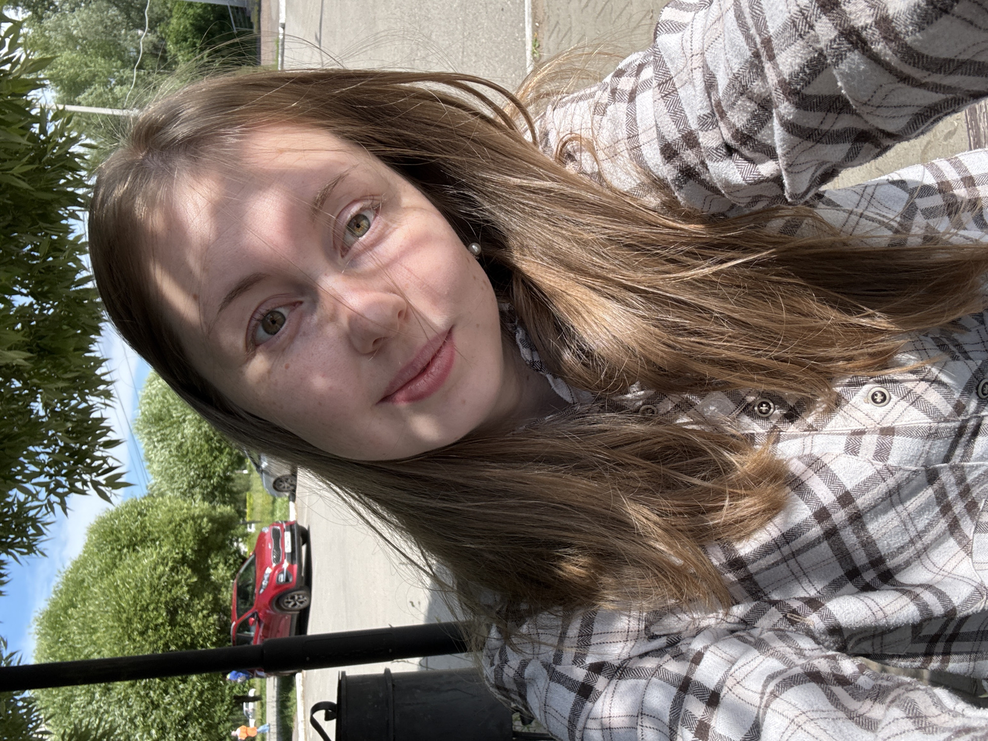 This is a close-up, low-angle selfie of a young woman with light brown hair and green eyes. She is wearing a plaid shirt in shades of white and brown. The photo appears to be taken outdoors on a sunny day, as evidenced by the strong sunlight and shadows on her face and the bright sky visible in the background. Trees and a red car are also visible in the background, suggesting a suburban or possibly rural setting. The woman has a gentle smile, and her expression conveys a sense of warmth and happiness. There is no visible text in the image.