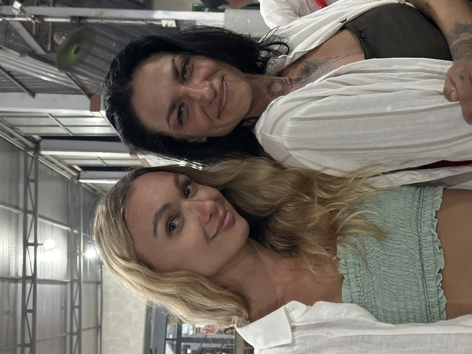 The image depicts two women smiling directly at the camera in an indoor setting within Phu Thuy Ward, Vietnam.
The woman on the left has long, wavy blonde hair with lighter highlights and blue eyes. She is smiling with a slight head tilt. She wears an unbuttoned white collared shirt over a light green, strapless, ruched bandeau top. Her skin tone is fair.
The woman on the right has dark, shoulder-length hair and dark eyes, smiling broadly. She exhibits a circular, sun-like tattoo on her neck/throat and a larger, intricate tattoo on her upper chest, possibly an eye or abstract design. A mandala-like tattoo is visible on her left forearm. She is wearing an unbuttoned white collared shirt with thin vertical light brown stripes, over a dark olive green ribbed tank top. Her skin tone appears slightly tanner than the other woman.
Both women are positioned in close proximity. The background reveals an industrial-style ceiling with exposed metal trusses, corrugated panels, and multiple bare light bulbs. On the right side of the background, a green industrial fan or light fixture is visible. The environment suggests a large, open public space, possibly a market or casual dining area, with diffused artificial lighting.