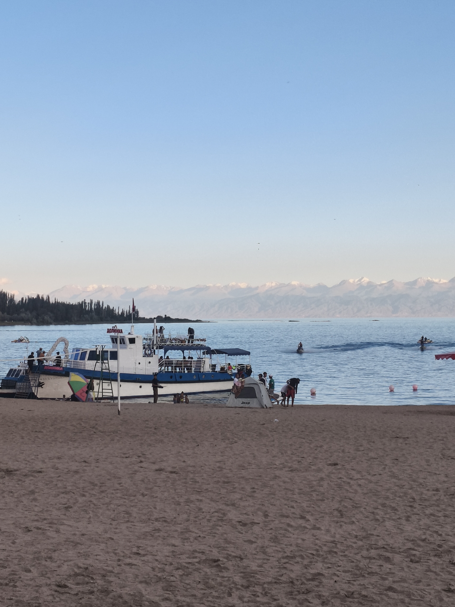 This image depicts a serene beach scene on the shores of Issyk-Kul Lake, near the city of Bayet, Kyrgyzstan. In the foreground, a sandy beach stretches out, with a few scattered objects and people. A large white boat with blue trim is moored near the shore, with several individuals visible on its deck and the surrounding area. One person stands on the beach next to the boat. Colorful beach umbrellas are visible, one of which is set up on the sand.
The middle ground features the calm, blue waters of Issyk-Kul Lake. Several people are enjoying water activities; some are on jet skis, creating wakes in the water, while others appear to be swimming or wading. Buoys are scattered in the lake, marking a designated swimming area.
In the background, a dense line of dark green coniferous trees borders the lake, backed by a majestic range of snow-capped mountains that stretch across the horizon. The sky is a clear, light blue, indicative of fair weather. The overall atmosphere is one of leisure and natural beauty.
