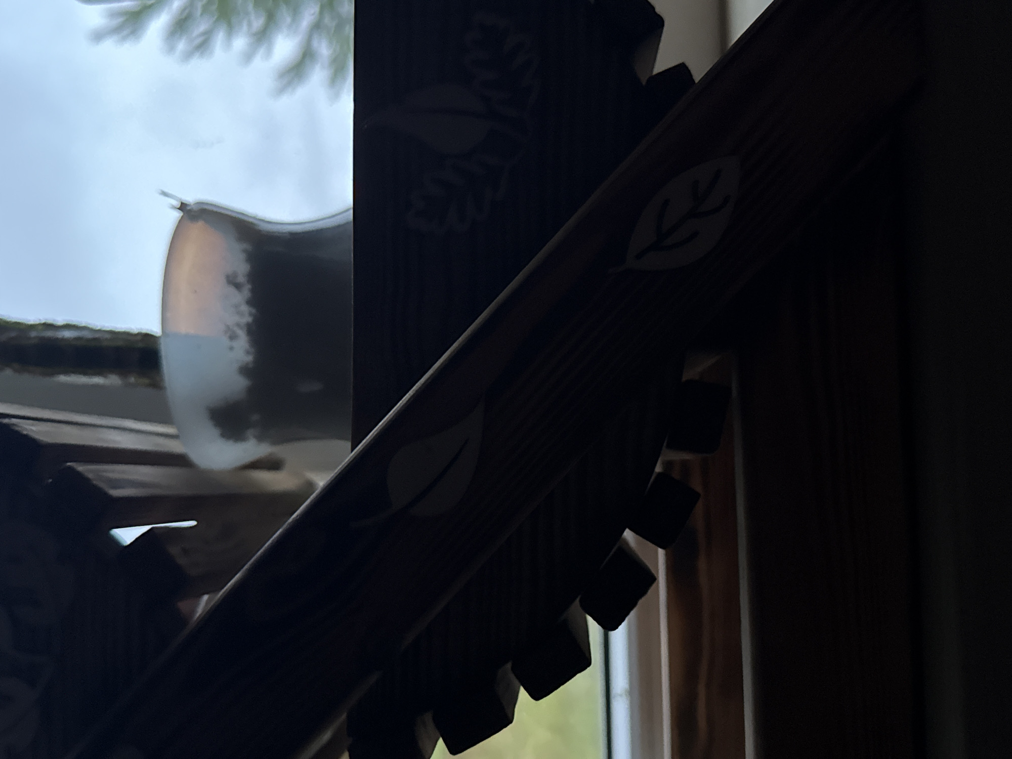 The image shows a close-up, angled view of dark wooden decorative elements, possibly part of a shelf or stand, with etched leaf patterns. These elements are positioned in front of a window with a cloudy sky visible outside, and a blurred white vase. The lighting is dim and casts shadows, making it difficult to discern finer details. The scene appears to be indoors, and while specific details about the location in Galinė, Lithuania are not directly visible, the overall aesthetic suggests a domestic or potentially a craft-oriented setting. There are no people or explicit actions depicted.