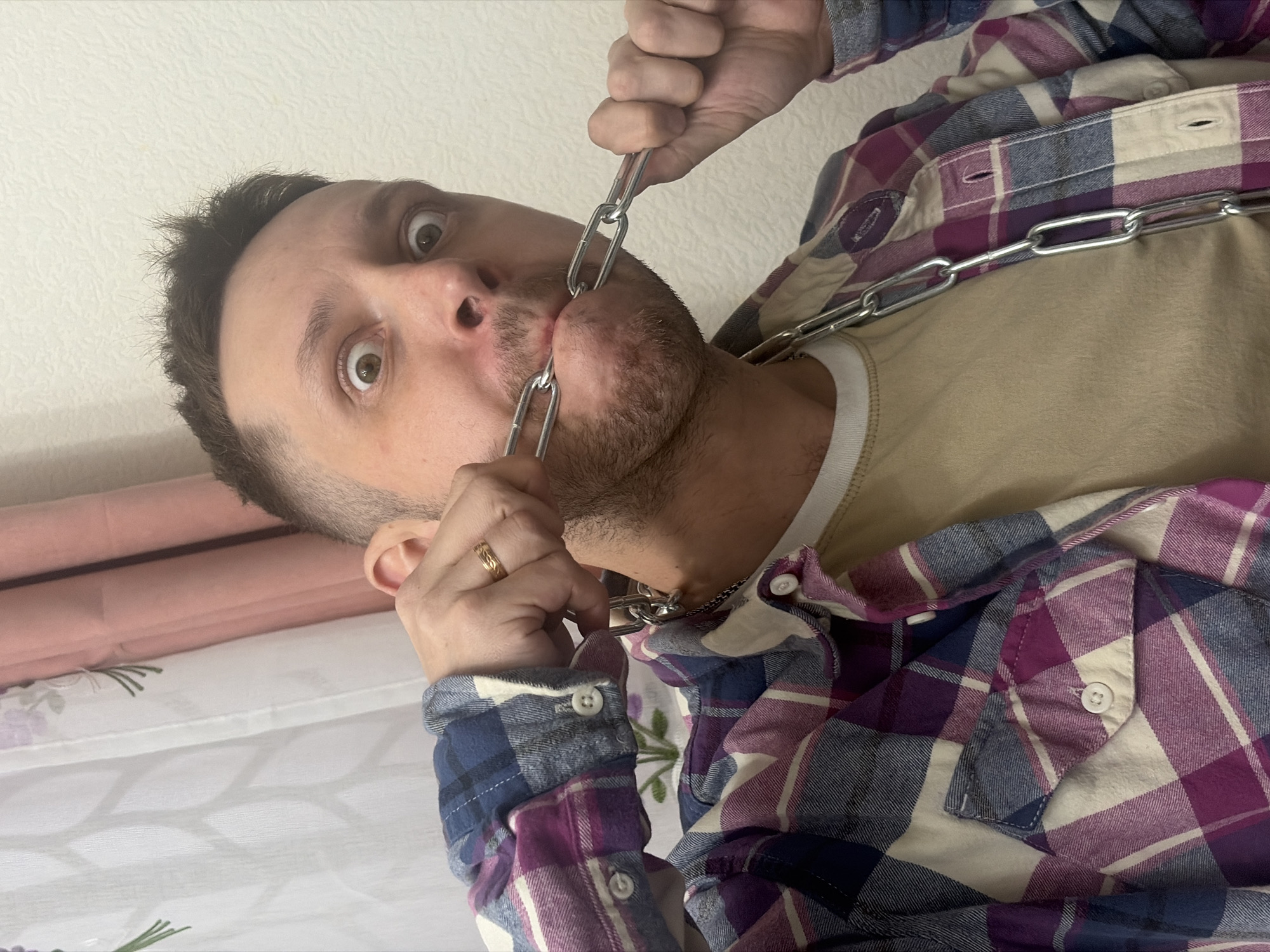 A light-skinned man is positioned off-center in the image, tilting his head backward and opening his mouth to thread a silver chain through it. He wears a plaid shirt in shades of blue, purple, and cream, over a tan undershirt. A gold ring is visible on his finger. The background consists of a white wall, with hints of a pink object and a white surface with floral patterns in the lower left corner. There is no evidence to determine if the location is in Saint Petersburg, Russia.