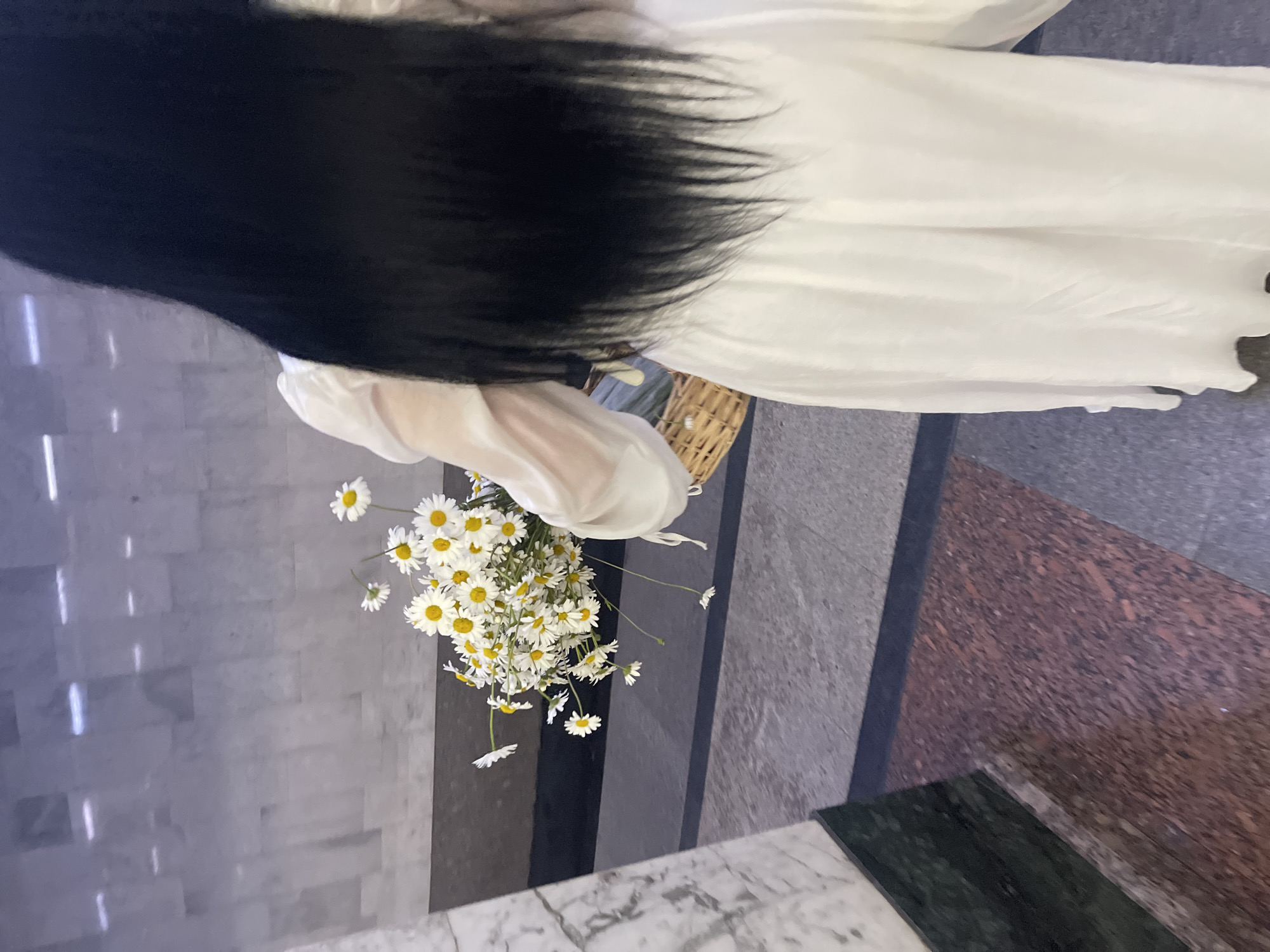 A person is depicted from behind, centered in the frame, walking away from the viewer. The individual has long, straight, black hair that extends to the mid-back. They are wearing a flowing, long-sleeved white garment, possibly a dress or a blouse and skirt combination, made of a light, semi-sheer fabric.
In the person's left arm, a voluminous bouquet of white daisies with prominent yellow centers is held. Several individual daisy stems dangle below the main bunch. A light brown, woven basket with a handle is carried on the person's right arm; its contents are obscured.
The immediate environment is an indoor public space. The background features tall, polished light grey marble walls with faint darker veining and horizontal panel lines. Multiple bright, linear reflections, consistent with overhead artificial lighting, are visible on the marble surface. To the left, a large vertical column, also clad in white marble, has a dark green marble baseboard. The floor exhibits a distinct pattern of materials: a section of dark grey, possibly granite or terrazzo, runs horizontally, separated by a thin black strip from a reddish-brown speckled terrazzo or granite section in the foreground and to the right. Another black strip separates the dark grey floor from the marble column base. The overall architectural style, characterized by grand use of marble and patterned flooring, is consistent with interiors found in the Moscow Metro, Russia.