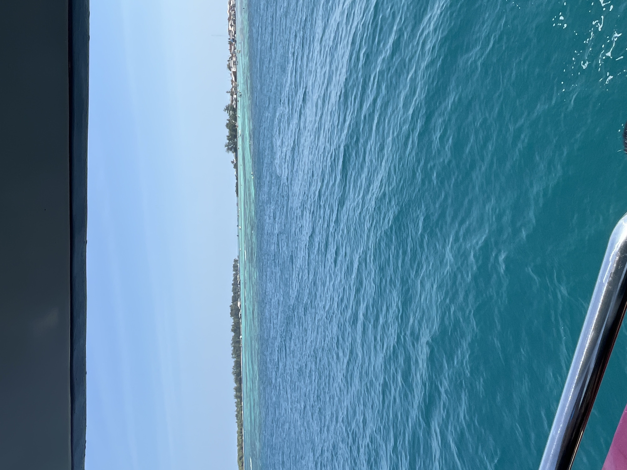 A wide-angle, eye-level shot captures a vibrant seascape off the coast of Karimunjawa, Indonesia. The foreground is dominated by the deep, rippling blue waters of the ocean, with a metallic railing visible along the bottom left edge of the frame. In the mid-ground, a shallow, turquoise lagoon separates the open sea from a long, narrow island. The island is densely populated with a variety of buildings, mostly white with red or brown roofs, nestled among lush green trees. Numerous small boats are scattered across the lagoon and near the shore, suggesting a bustling coastal community or a popular tourist destination. The sky above is a clear, bright blue, unblemished by clouds. The overall impression is one of a tropical paradise, serene yet active.