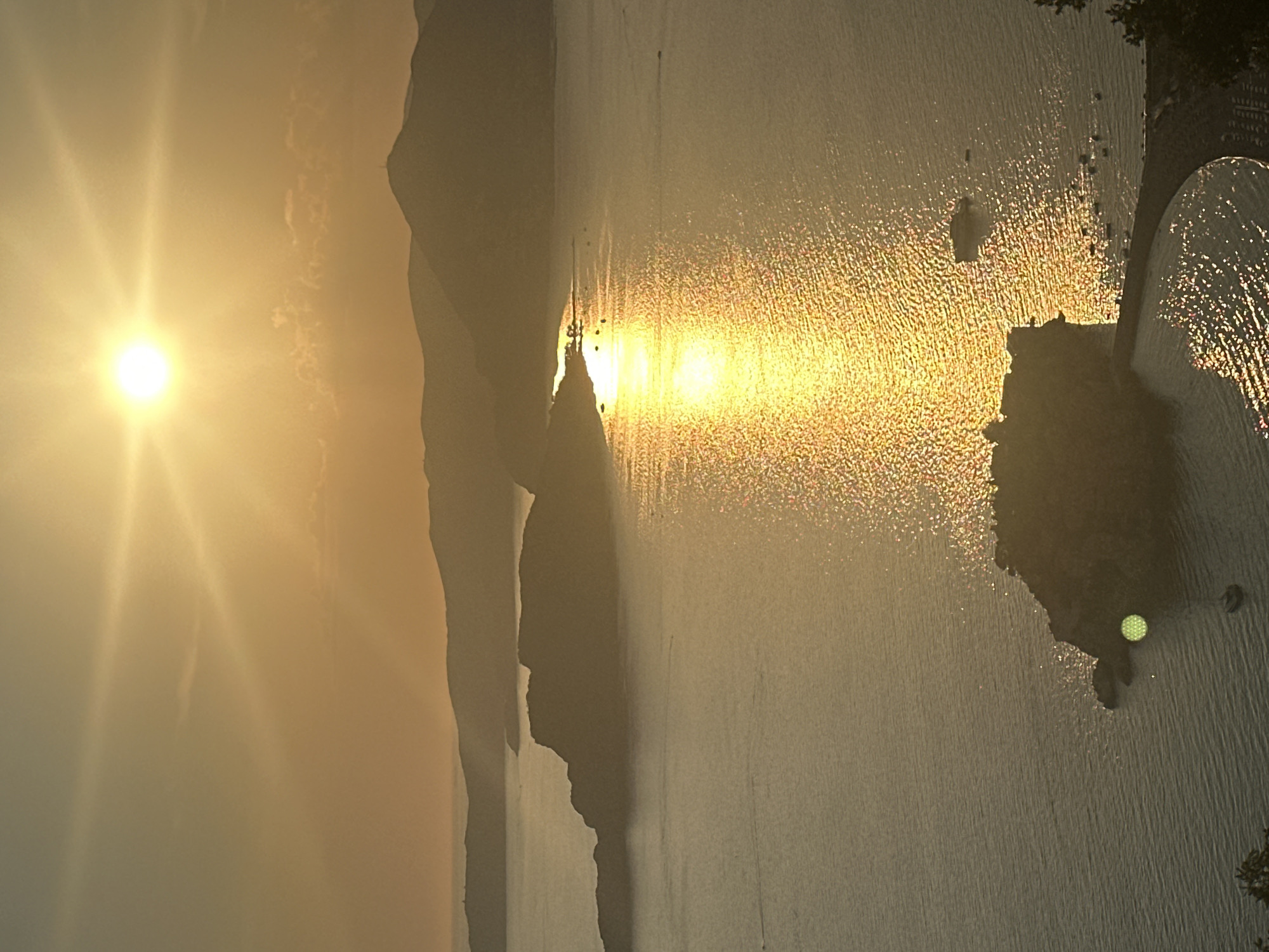 The image depicts a sunset over the sea near Sveti Stefan, Montenegro. The sun is a bright, intense orb in the upper center of the frame, casting a golden, shimmering reflection across the water. Silhouetted islands and coastlines with rolling hills form the background, their shapes softened by the haze of the setting sun. In the foreground, a small island, likely Sveti Stefan itself, is visible, connected to the mainland by a narrow causeway. A few boats are scattered on the water, and the surface of the sea is textured with ripples and the bright glare of the sunlight. The overall atmosphere is serene and picturesque, emphasizing the natural beauty of the coastal landscape at dusk.