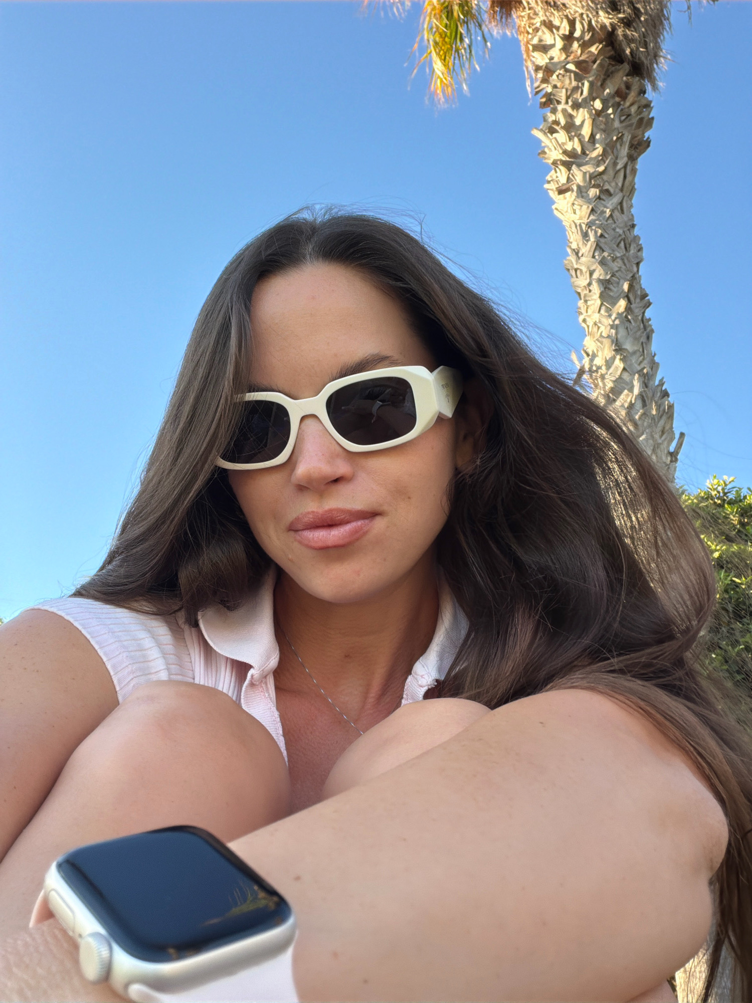 A woman is captured in a selfie outdoors, with a bright blue sky serving as the background, and a palm tree trunk and foliage visible on the right. She is wearing a light pink collared shirt, white rectangular sunglasses, and a smartwatch on her left wrist. Her long, dark brown hair is spread around her shoulders. Her knees are drawn up towards her chest, and her arms are around them. The lighting suggests it is daytime. The scene is consistent with an outdoor location in Salou, Spain, known for its beaches and palm trees.