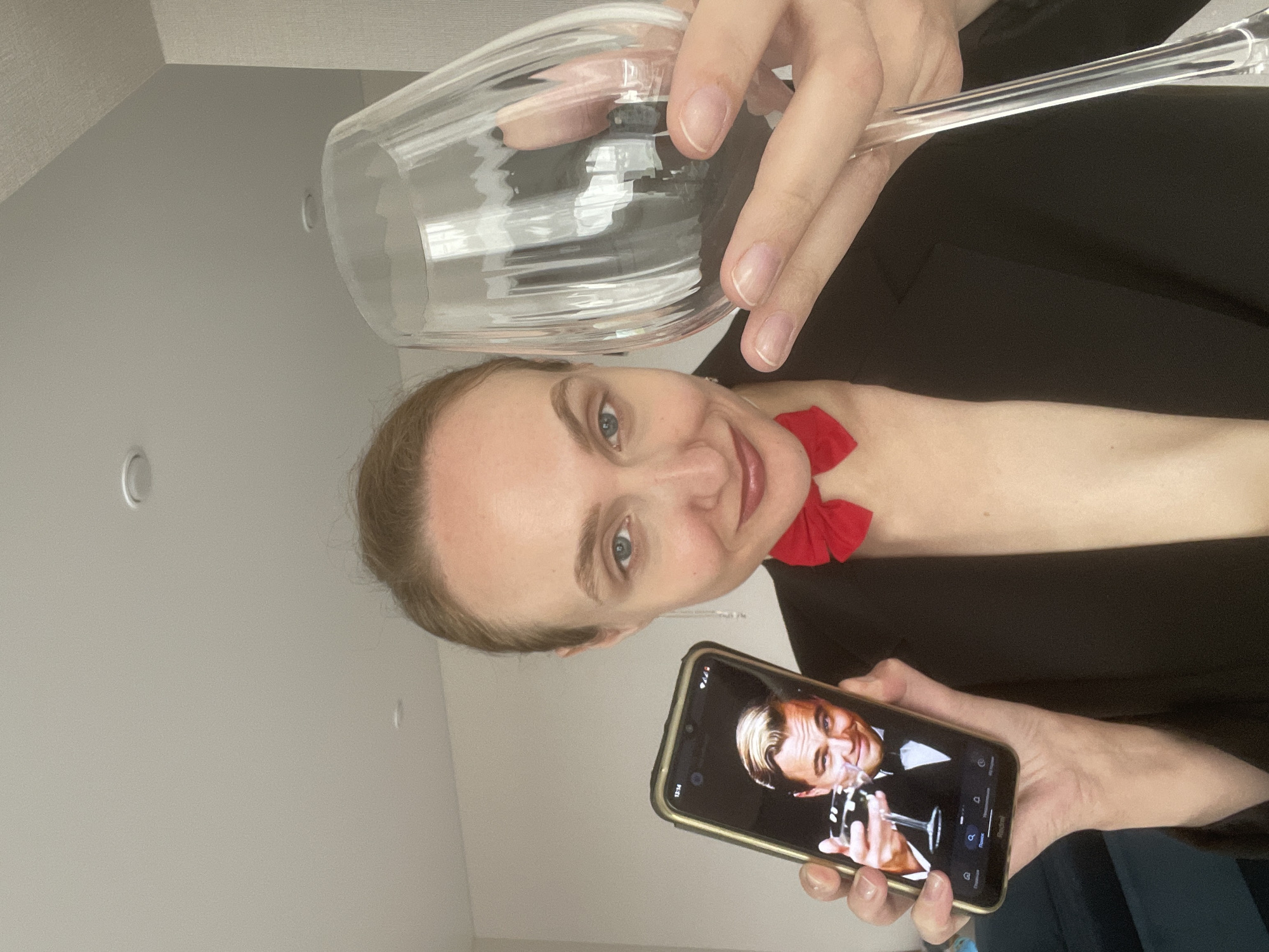 A fair-skinned woman in a black suit and red bow tie is holding a wine glass in her left hand and a smartphone in her right hand. The smartphone screen displays an image of Leonardo DiCaprio in a black suit and bow tie, also holding a wine glass. The background is a plain, light-colored wall with a ceiling light fixture. The location is unknown and cannot be determined from the image. The city of Подшивалово, Russia is not indicated in any way in the image.