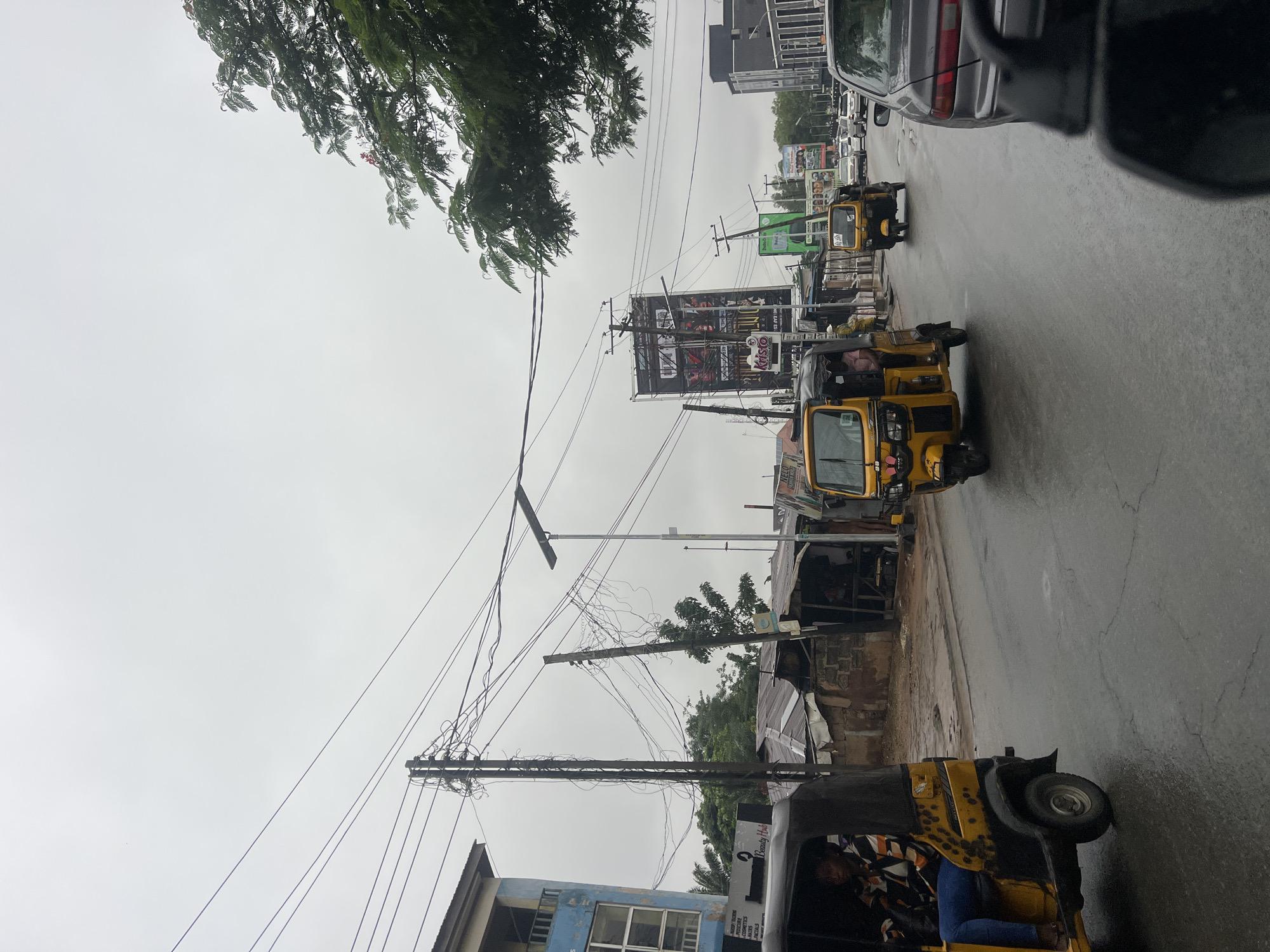 The image depicts a street scene in Onitsha, Nigeria, under an overcast sky. The road is wet, suggesting recent rain. Several yellow auto-rickshaws, commonly known as "keke" or "marwa," are present, with one in the foreground on the left and another further down the street on the right. A dark-colored sedan is also visible in the right lane, with its rear end facing the camera.
On the left side of the street, a multi-story blue building stands, with a sign that appears to read "Beauty Hub." Adjacent to it are low-rise structures with corrugated metal roofs, indicative of informal market stalls or small businesses. Prominent electrical poles with a dense network of overhead wires are scattered throughout the scene, a common sight in urban Nigerian environments.
In the background, larger billboards with advertisements are visible, adding to the urban landscape. Green trees with foliage are interspersed with the buildings and infrastructure. The overall atmosphere is one of a bustling street in a Nigerian city, with everyday traffic and commercial activities taking place.