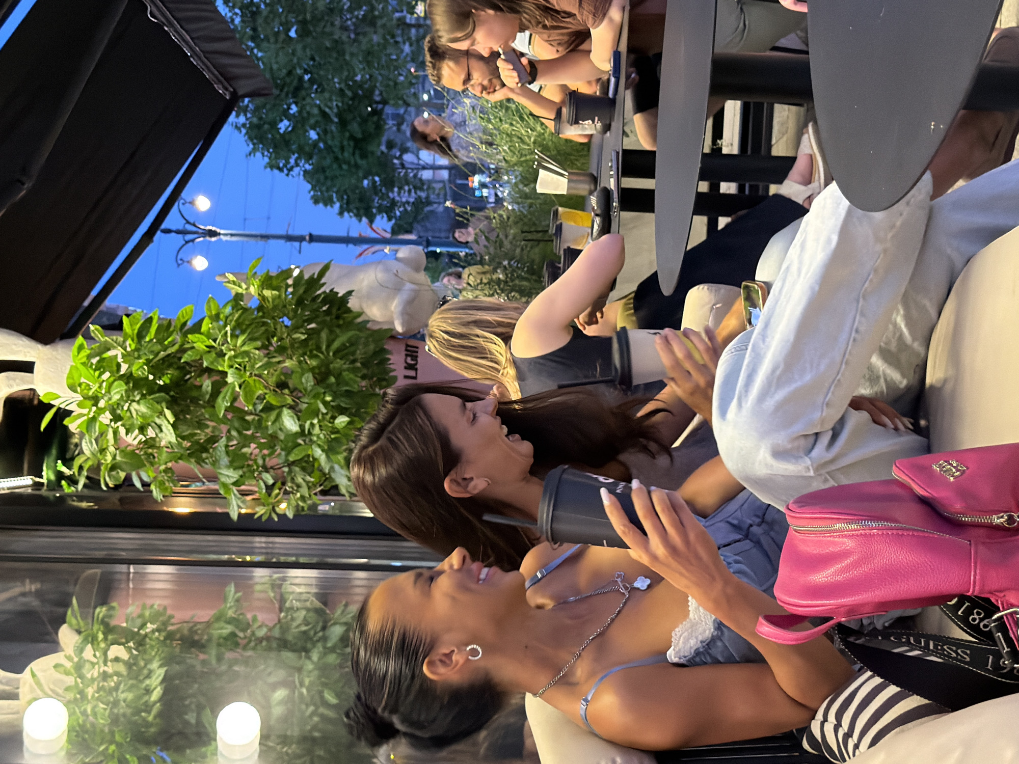 Two women are seated at a table at an outdoor cafe in Novaya Usman', Russia. The woman on the left has her hair in a bun and is wearing a blue camisole with lace trim and a silver necklace. She is holding a black cup and laughing. The woman on the right has long brown hair and is wearing a grey top. She is also holding a black cup and smiling. In the background, other patrons are seated at tables, and there are plants and streetlights visible. The sky is a deep blue, suggesting it is evening. A pink backpack is on the seat next to the woman on the left.