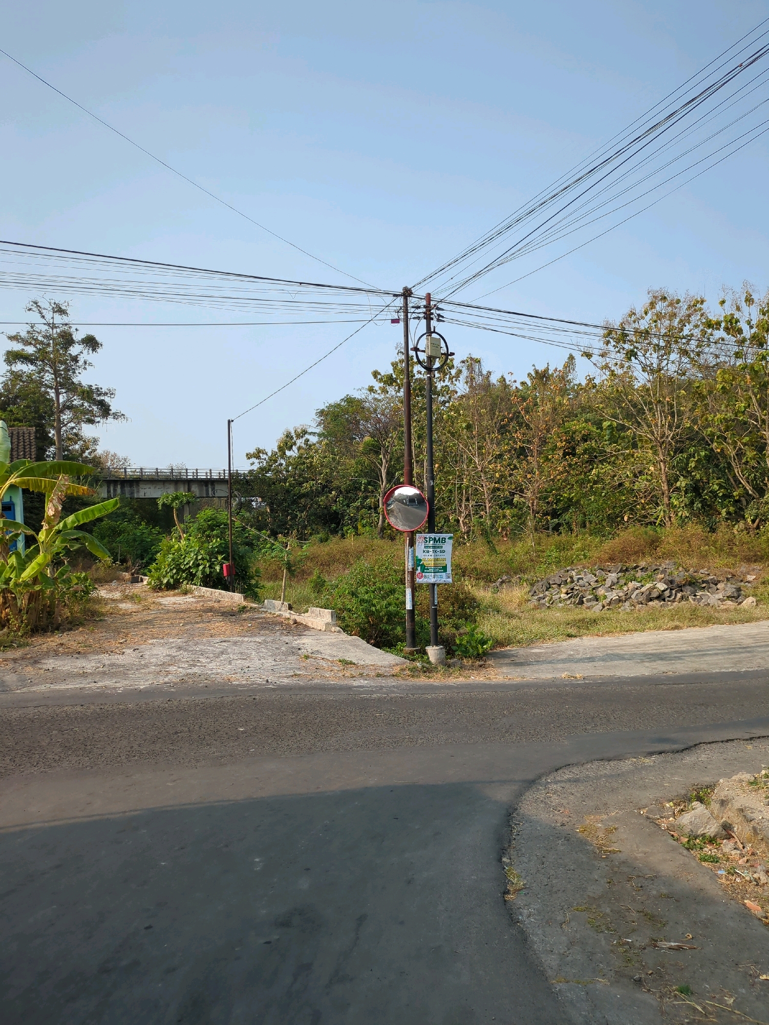 The image depicts a rural road intersection in Sindon, Indonesia, bathed in the bright light of a clear sky. A paved road curves into the foreground, transitioning to a more textured, sun-drenched surface with debris and sparse vegetation on the right. Overhead, a dense network of power lines crisscrosses the sky, supported by utility poles.
To the left of the road, a dirt path leads towards a concrete bridge structure visible in the distance, spanned by trees and lush green foliage. Banana plants with large leaves stand prominently on the left edge of the scene.
In the center, a utility pole stands tall, equipped with a round, convex mirror, likely for traffic visibility, and a sign with Indonesian text that reads "SPMB" and appears to be related to educational institutions. Another smaller red object is attached to the pole below the mirror. To the right of the pole, a pile of rough, grey stones sits on a grassy embankment. The overall environment is characterized by a mix of natural vegetation, man-made infrastructure, and a sense of rustic tranquility under the midday sun.