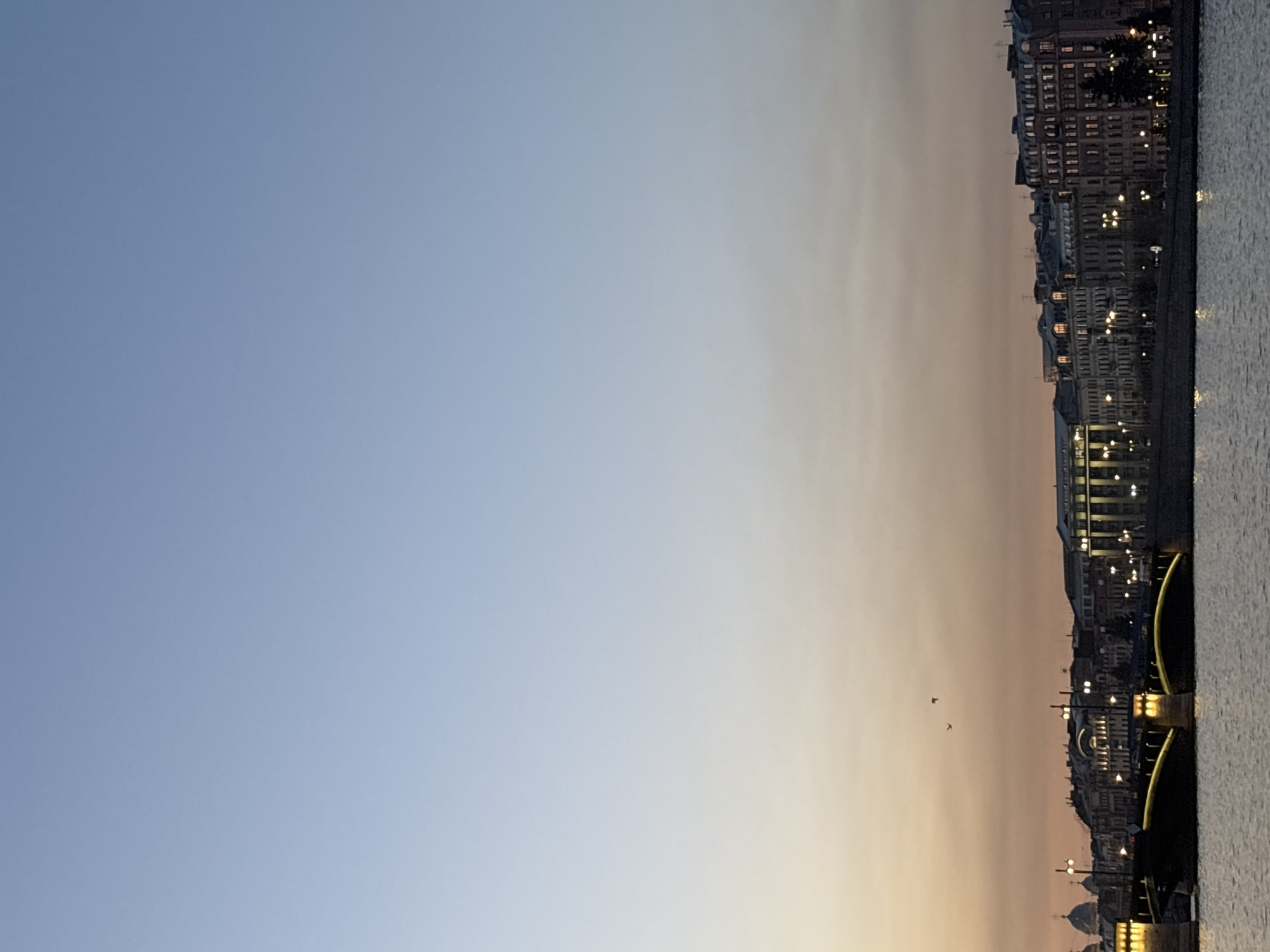 The image shows a cityscape at dusk, likely taken in Saint Petersburg, Russia. The right side of the image depicts a body of water, possibly the Neva River or a canal, adjacent to a row of buildings. The architecture suggests a classic Petersburg style. Lights are visible from the buildings and street lamps along the embankment. A bridge with golden arches spans over the body of water. The sky transitions from a clear blue on the left to a warm, light orange near the horizon. Two birds are visible in the distance near the horizon. The scene appears calm and serene.