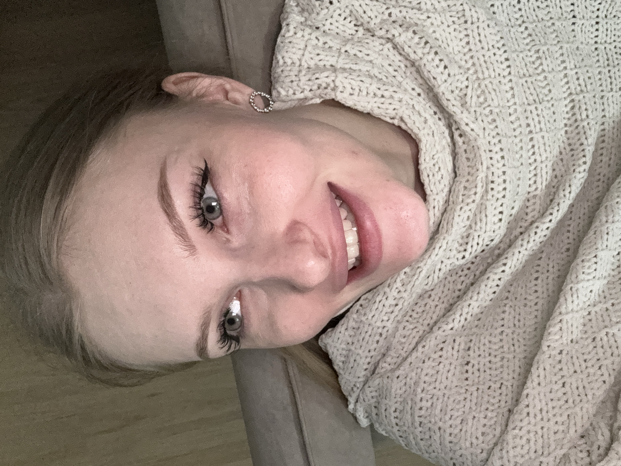 A young woman with fair skin, light hair, and gray eyes is looking up at the camera. She is wearing a light-colored knit sweater or shawl draped around her. She has a silver earring in her left ear. The background appears to be a room with a neutral-colored wall or furniture. The location is not discernible from the image.