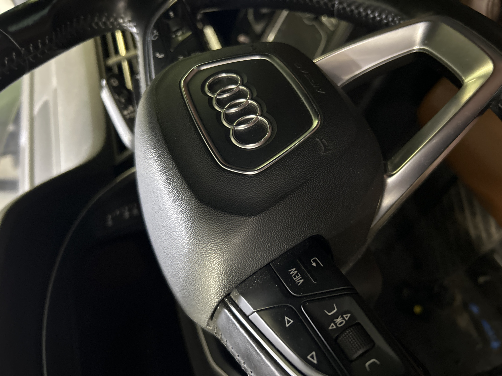 The image shows a close-up view of a black Audi steering wheel, featuring the iconic four-ring Audi logo in silver. The steering wheel has various control buttons, including directional arrows, an "OK" button, and a "VIEW" button. The car interior is dimly lit, suggesting it might be parked indoors, possibly in a garage or car dealership. The background is blurred, making it impossible to identify any specific city, including Tallinn, Estonia. There are no people or any discernible actions or interactions captured in this image.