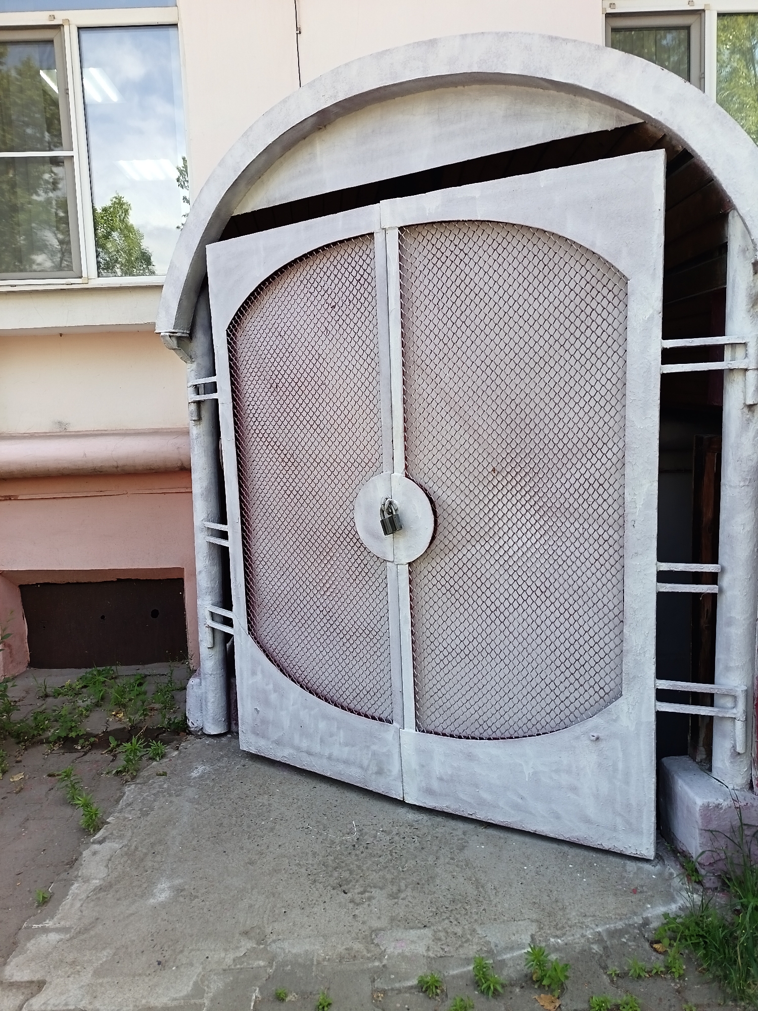 The image depicts an exterior view of a building entrance in Komsomolsk-on-Amur, Russia. A large, light grey metal gate with diamond-shaped mesh infill is partially open. The gate is secured with a padlock. The entrance is framed by a curved concrete arch. To the left of the gate, a section of the building facade is visible, painted in a pale pink color, with a window featuring reflections of the sky and trees. Below the window, a dark opening with a metal cover is present, and the ground in front of it is covered with sparse green vegetation. The ground surface is concrete, showing some cracks and wear. To the right of the gate, a vertical metal pipe and other architectural elements are visible. The scene appears to be an access point to a basement or service area. No people are present in the image.