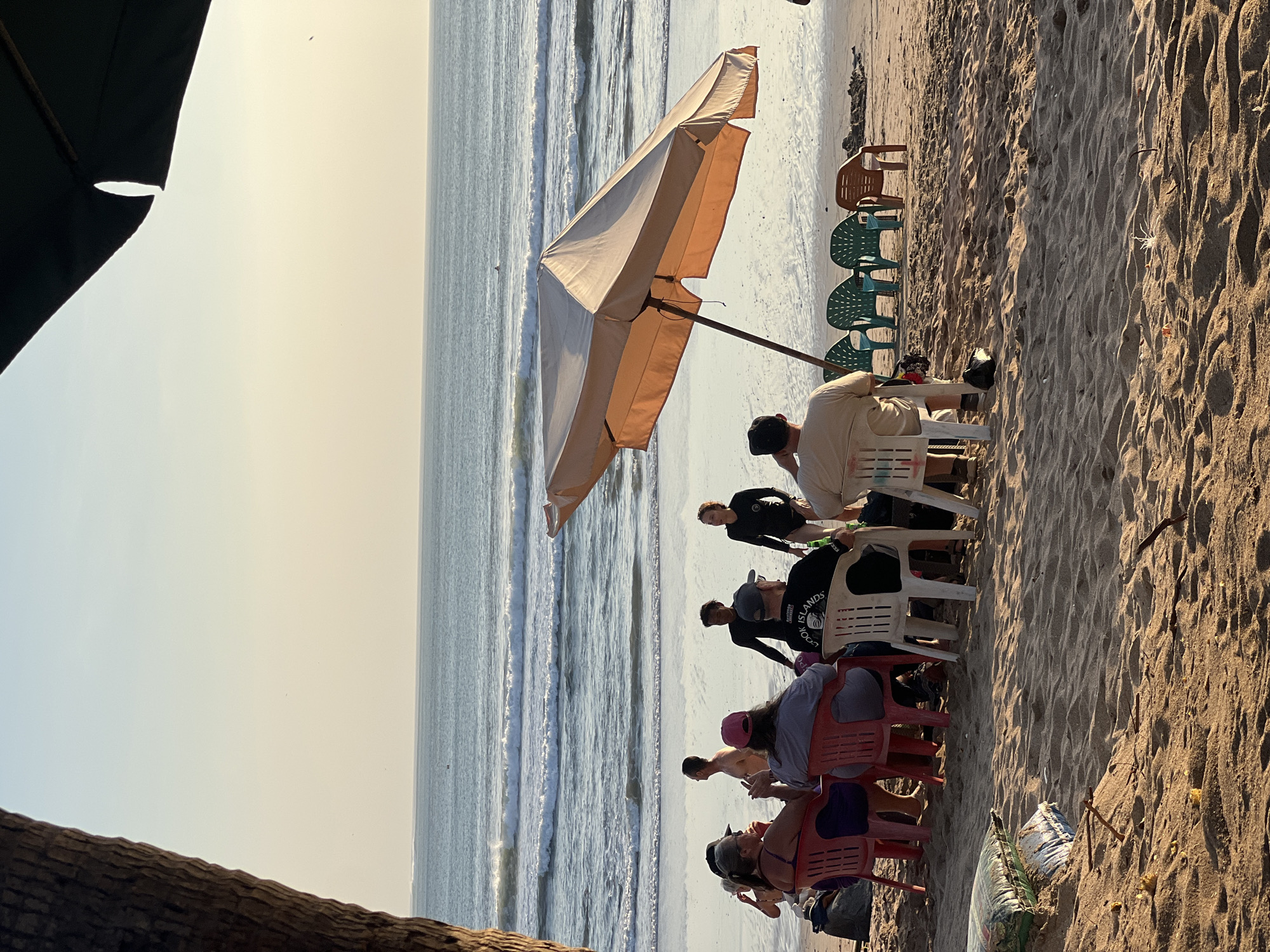 The image depicts a beach scene in Kuta, Indonesia, at sunset. A group of approximately ten people are seated on plastic chairs on the sandy shore, facing the ocean. The ocean has moderate waves, and a lone surfer is visible in the distance. A large, tan beach umbrella shades some of the chairs. The sand is littered with small pieces of driftwood and debris. The sky is a pale orange and blue, indicating the time of day. The composition is framed by a tree trunk on the left and the edge of a dark umbrella on the right.