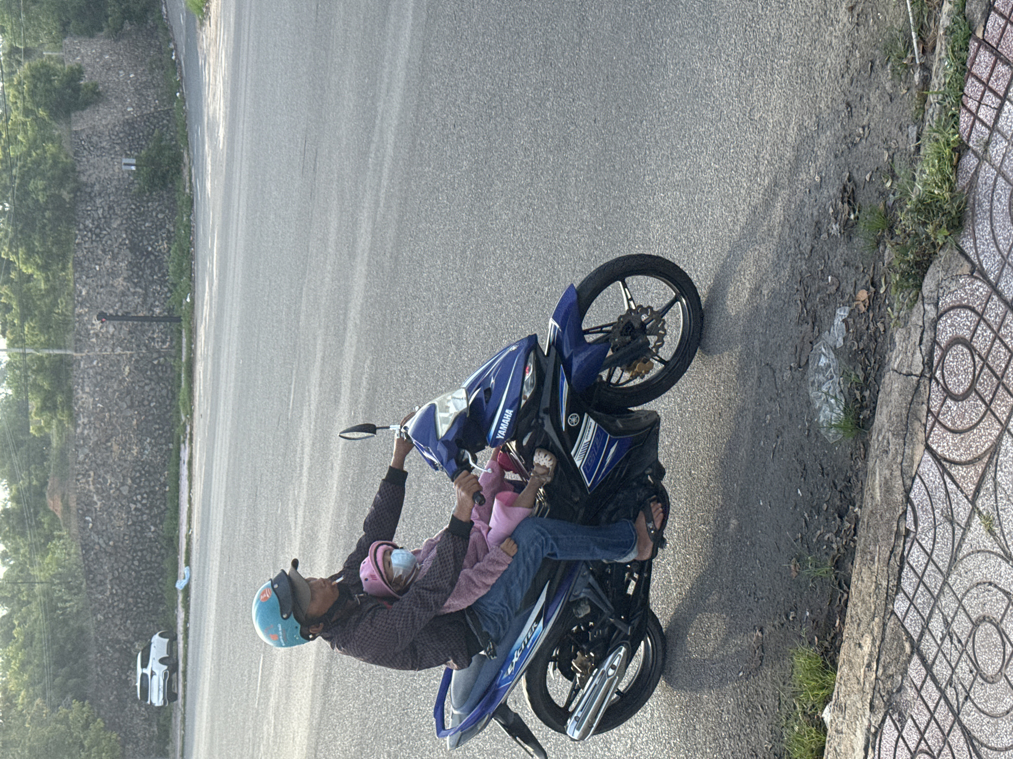 A person wearing a blue helmet and dark patterned shirt is riding a blue and black Yamaha motorcycle on a paved road. They are carrying a young child in front of them, who is wearing a pink outfit and a light blue face mask. The child also has a pink hat on. The motorcycle has the words "EX-ITER" and "GTR" visible on its body. The rider is wearing blue jeans and dark flip-flops. The scene appears to be in Mũi Né, Vietnam, with a stone wall and trees in the background, and a tiled sidewalk along the side of the road. The sun is casting a shadow of the motorcycle and riders on the road.