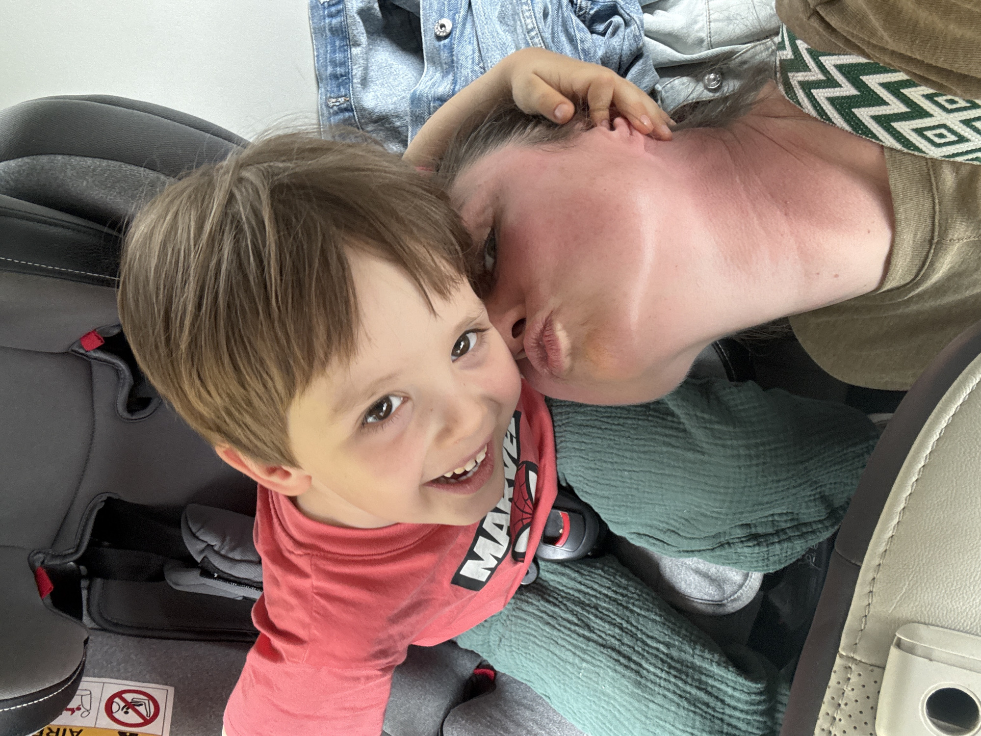 A photograph taken from a high angle shows a child and an adult, likely in a vehicle. The child is in a car seat, wearing a red t-shirt with white and black lettering. The child is looking up and smiling at the camera. The adult is leaning in, with their face close to the child's face, as if giving a kiss. The adult is wearing a green top with a white geometric pattern. The seat in the background is gray, and there's a blue denim jacket or shirt. The car's interior is visible. It is not possible to ascertain the location as Meljine, Montenegro, from this image.