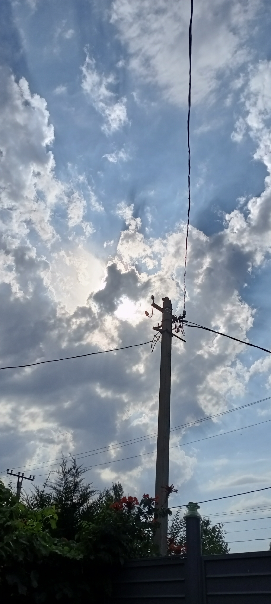 The image shows a utility pole in the city of Sokuluk, Kyrgyzstan, set against a sky filled with dramatic clouds. The sun is partially obscured by the clouds, casting rays of light that create a luminous effect. The utility pole itself is made of concrete and supports electrical wires and insulators. To the left and bottom of the pole, there is lush green foliage and some red flowers, suggesting a natural setting within the urban environment. In the background, other utility poles and wires are visible, indicating a residential or semi-urban area. The fence in the foreground is dark and appears to be made of metal or wood.