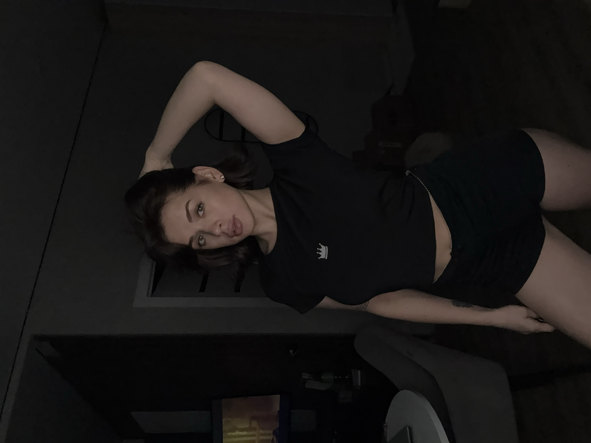 The image depicts a woman in a dark room, lying on her side. She is wearing a black t-shirt with a crown logo and black shorts. Her left arm is raised above her head. The room appears to be a living space with a chair and a television in the background. The exact location in Лобаново, Russia is not discernible from the image.